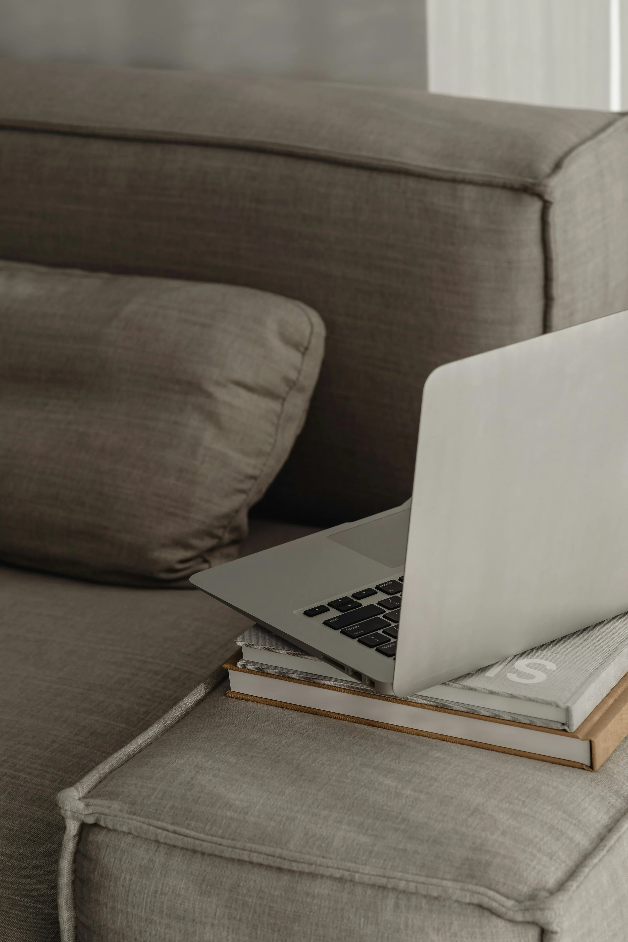 A silver laptop is on top of a stack of books on a gray upholstered ottoman. The background shows part of a gray sofa.