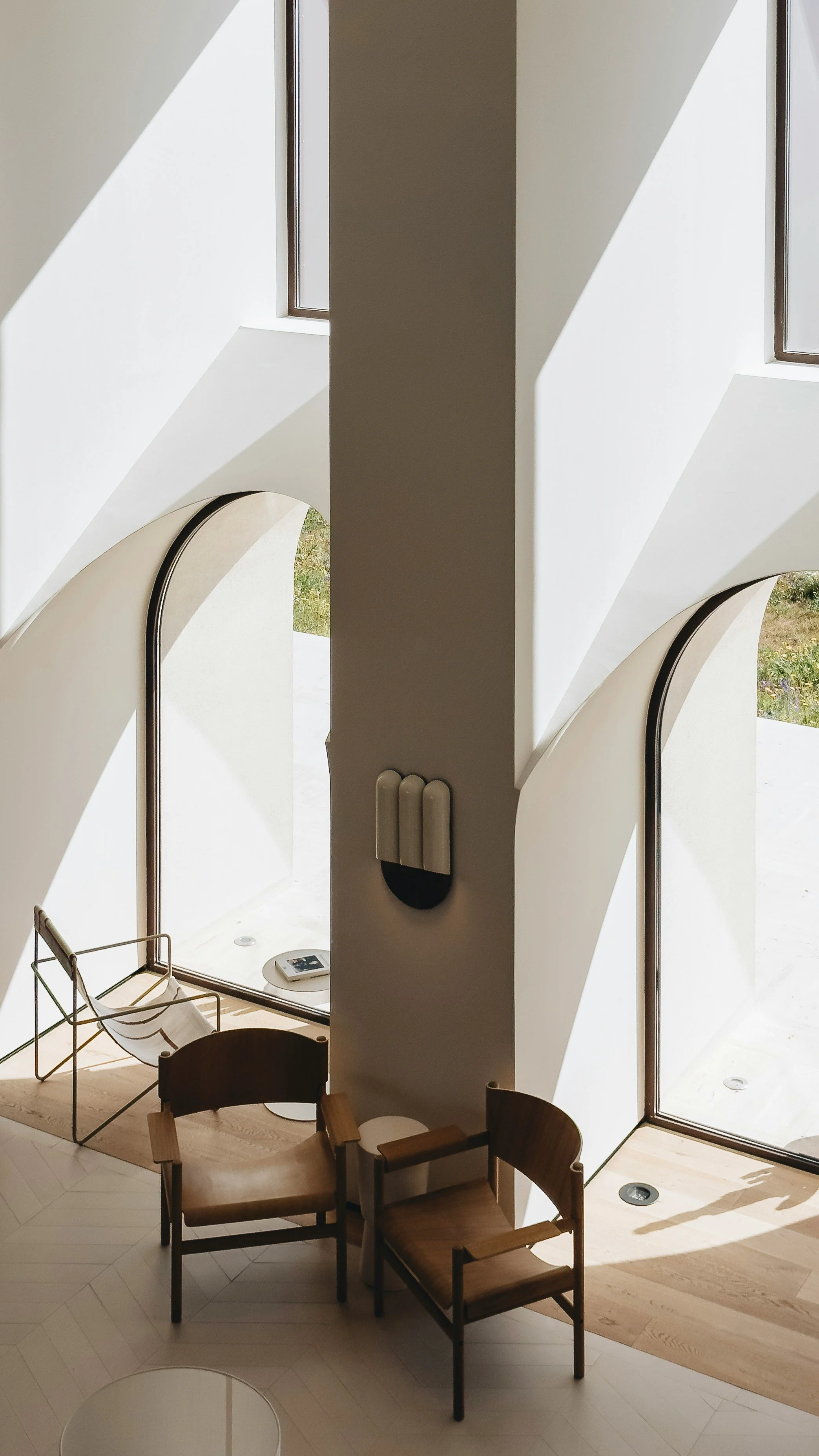 Interior of a modern building with large windows letting in natural light, white walls and curved architectural features. There are wooden chairs and a small table on a light-colored wooden floor.