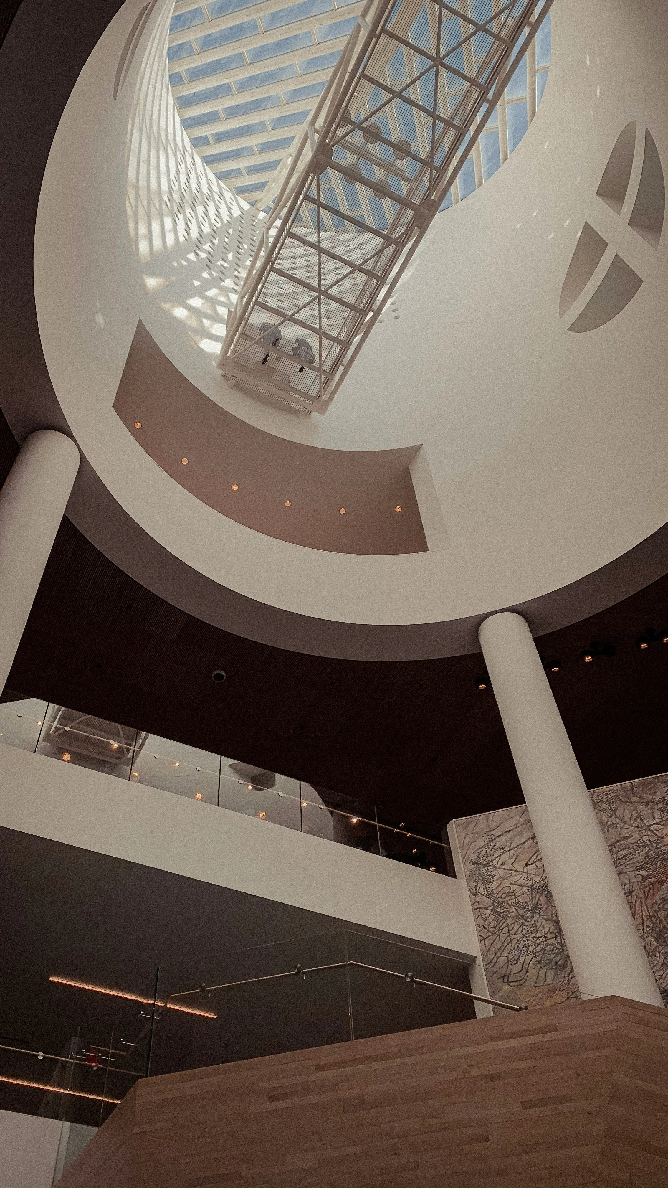Interior view of a modern building with a circular opening in the ceiling, showcasing a glass roof and metal staircase, with curved white walls, wooden accents, and architectural lighting.