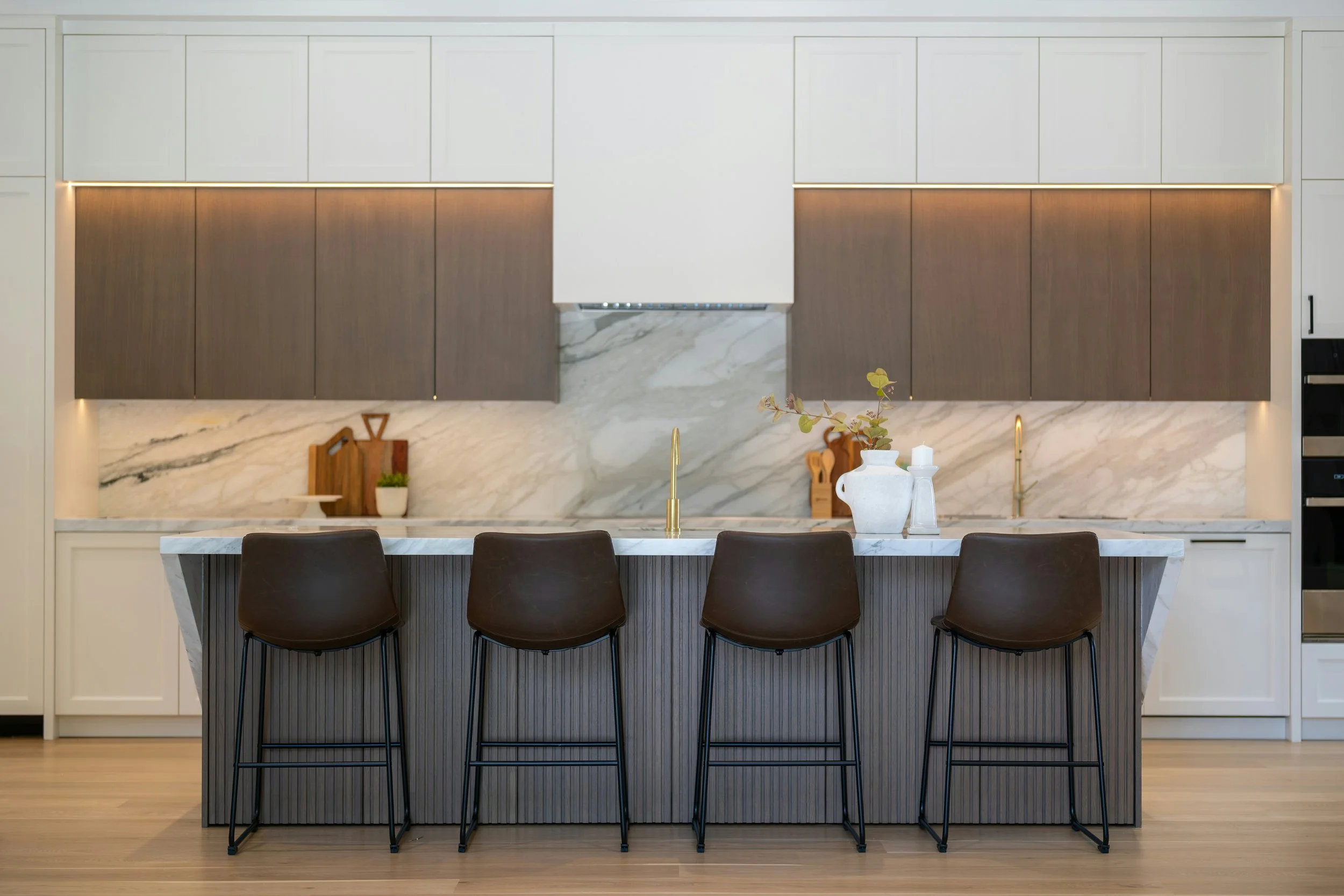 Modern kitchen with white and wood cabinets, marble backsplash, kitchen island with dark chairs, and decorative vases on the counter.