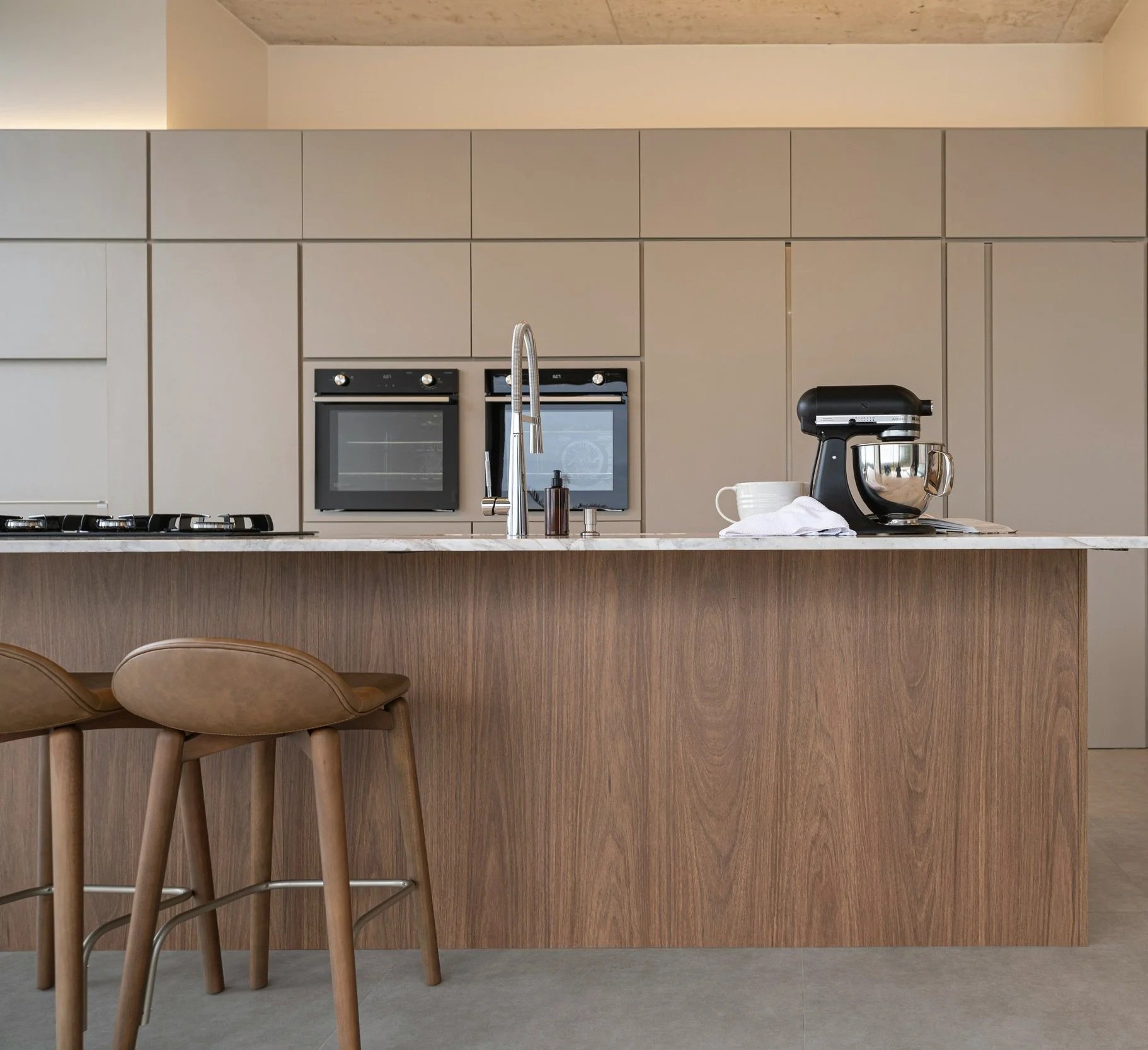 Modern kitchen with light beige cabinets, gray marble countertop, built-in black oven, stand mixer, white mug, and brown chairs with curved backs.