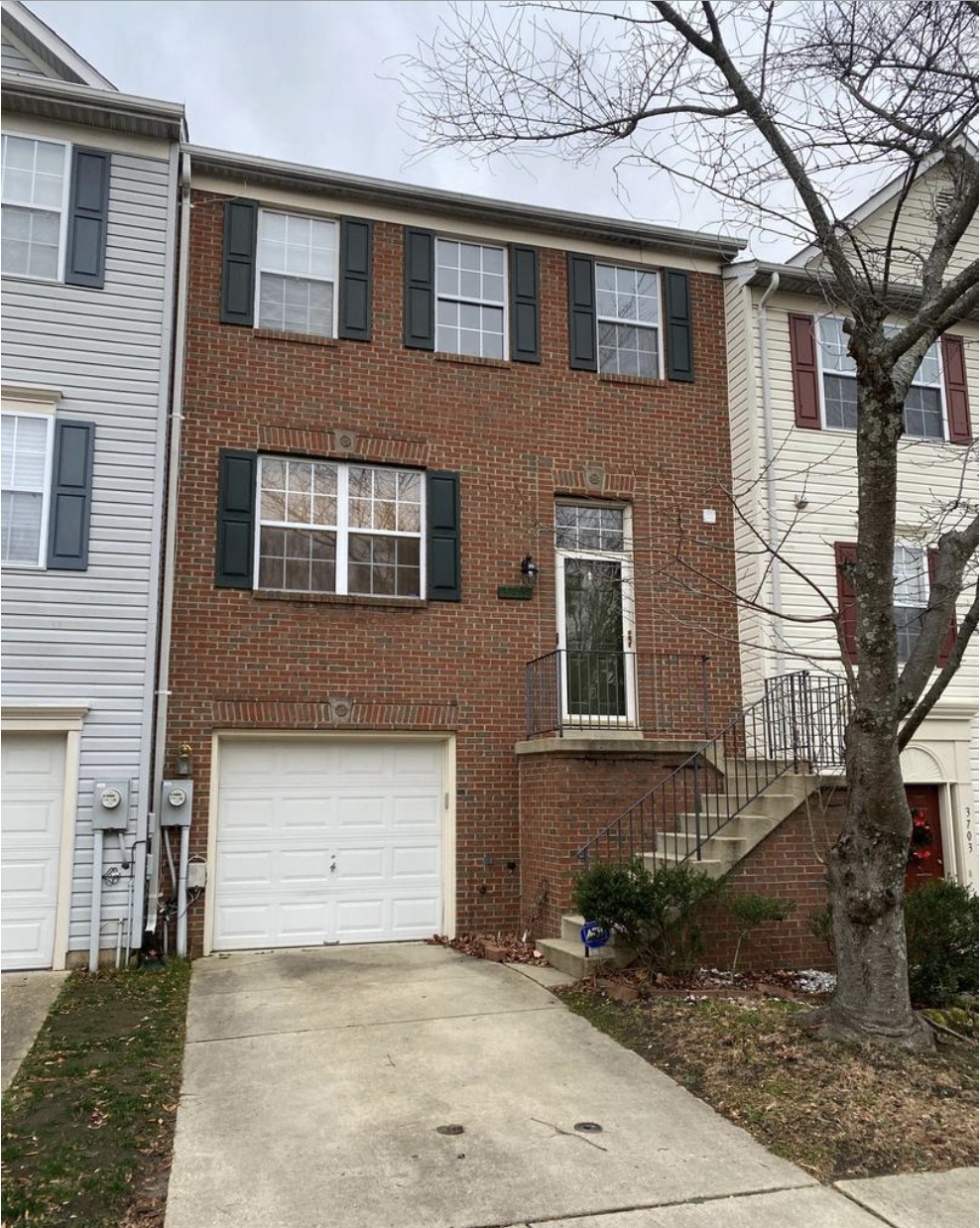 A three-story townhouse with a brick facade, black window shutters, and a white garage door. There are stairs leading up to the front door with a small landing and black railing. A tree with no leaves is in front of the house, and there are some bushes near the stairs.