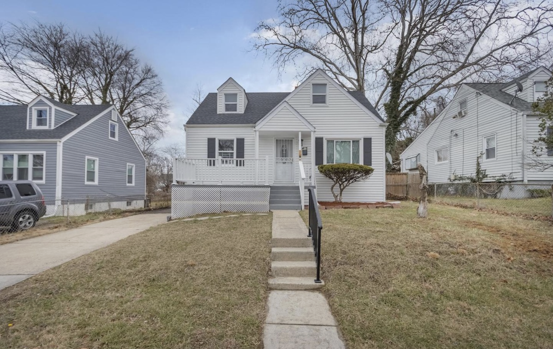White two-story house with a small front porch, black shutters, and a gabled roof, situated between two other houses. There are leafless trees in the yard and a concrete sidewalk leading to the front stairs, with a grassy lawn on either side.