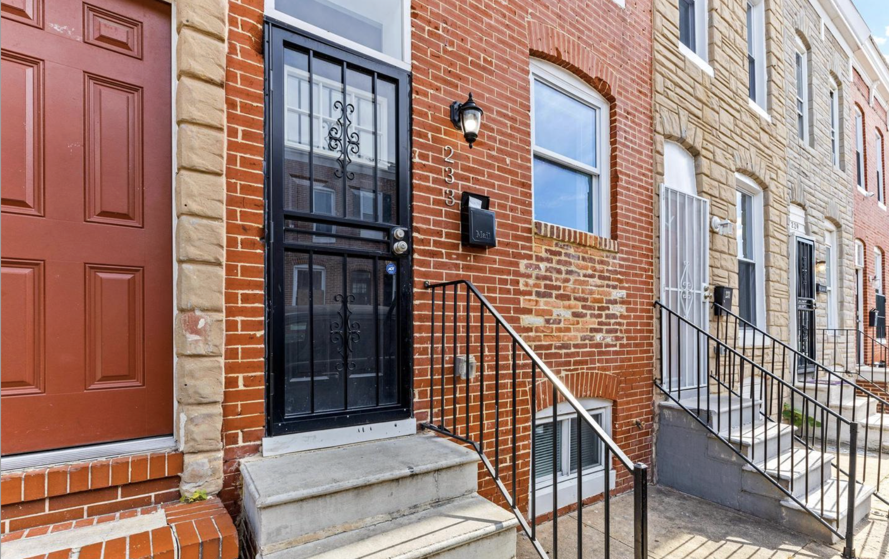 Row of brick townhouses with stairs leading to the front doors, black metal railings, and security screens on the doors. One door has a lamp next to it, and the houses have various window styles.