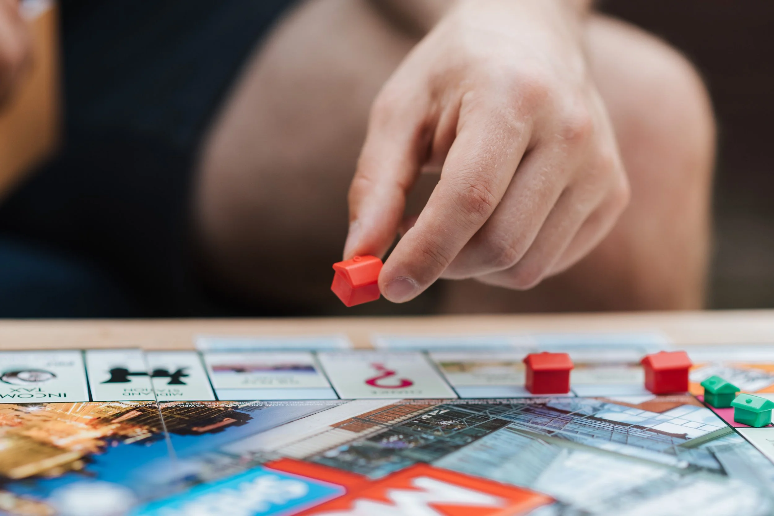A close-up of a person's hand moving a red game piece on a colorful board game with various illustrated cards and game components.