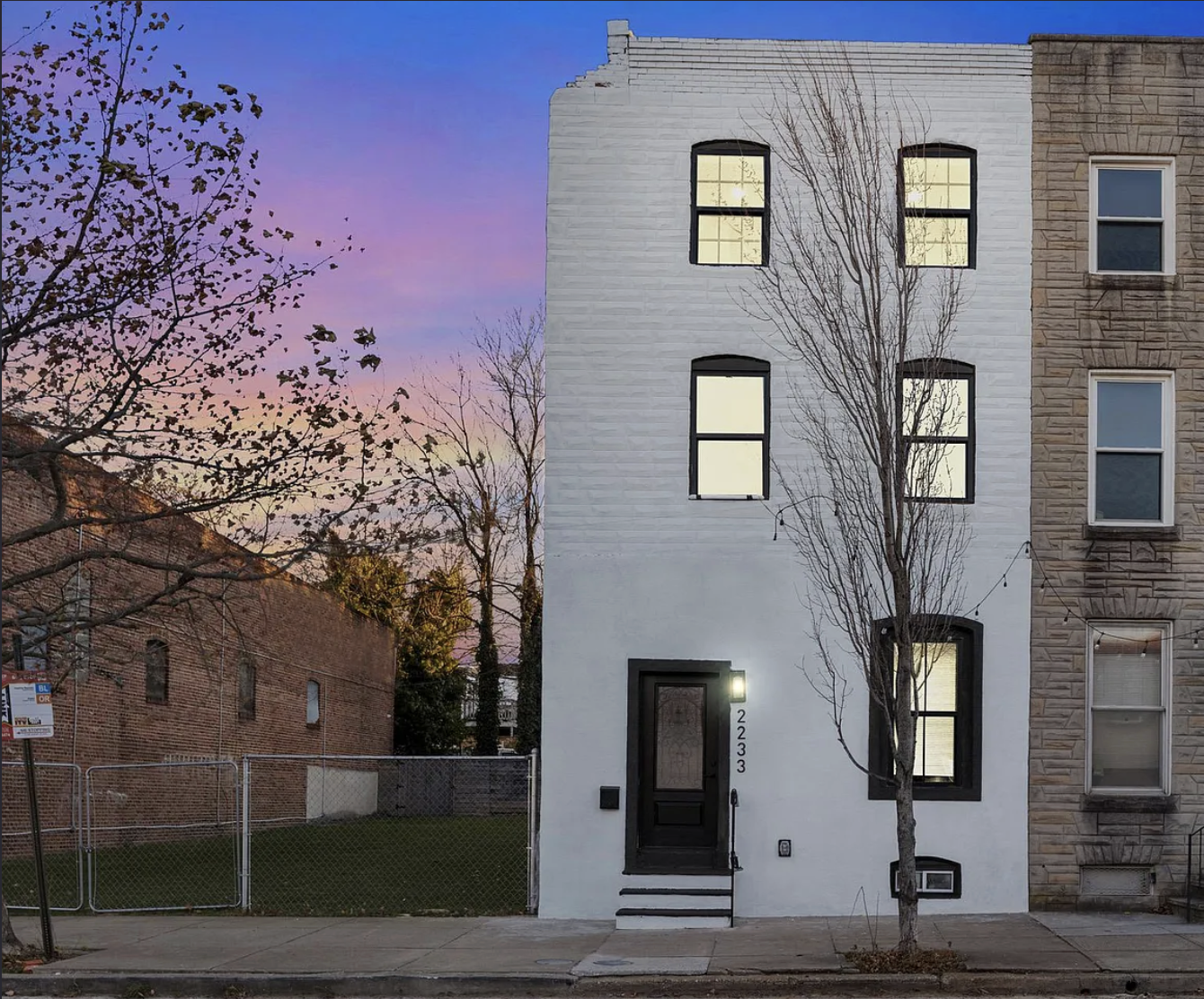 A narrow, white, three-story townhouse with black-framed windows and a black front door, located between two other buildings, with a bare tree in front, and a sunset sky in the background.
