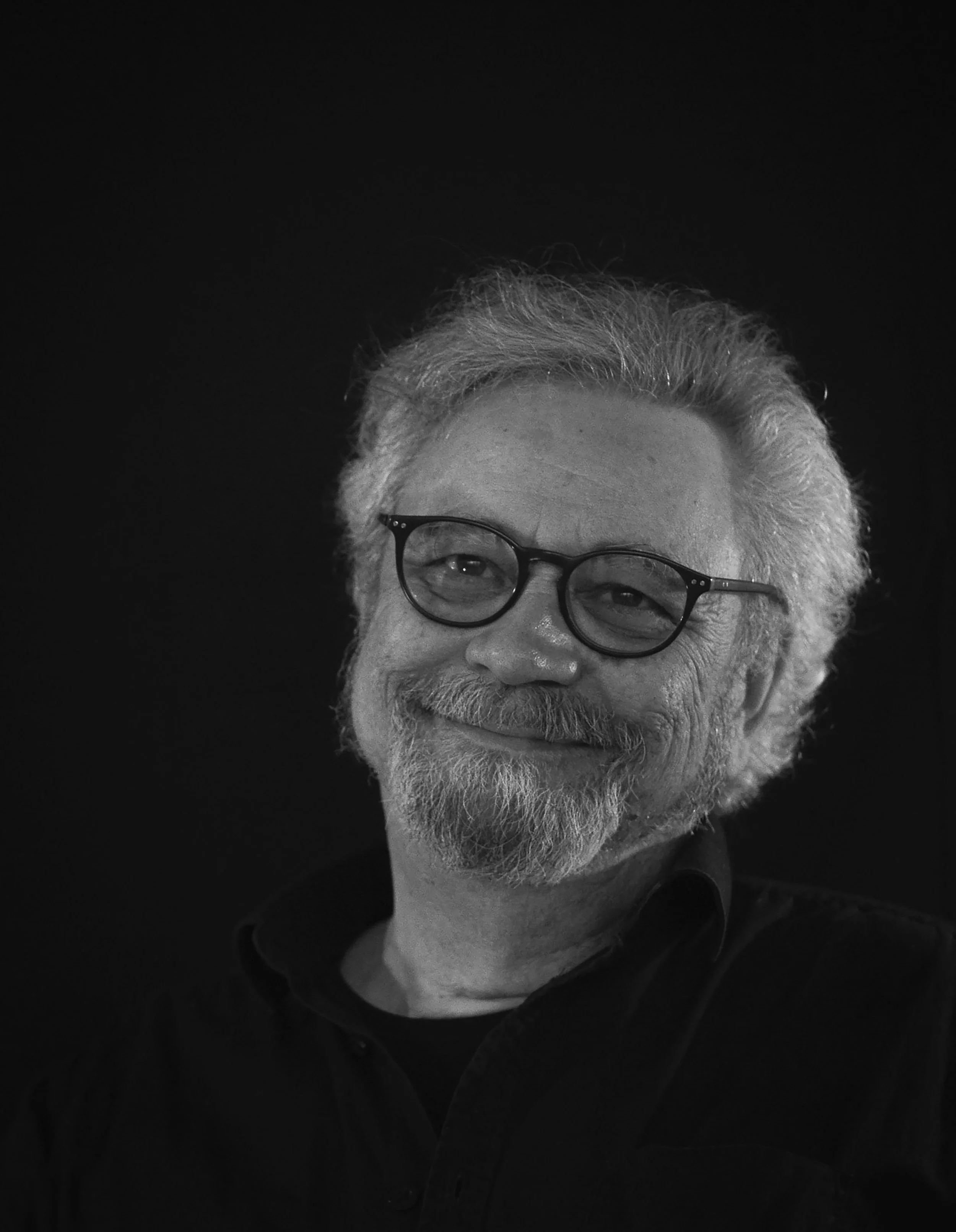 Portrait en noir et blanc d'un homme âgé avec des cheveux gris, une barbe blanche, portant des lunettes, avec un sourire.