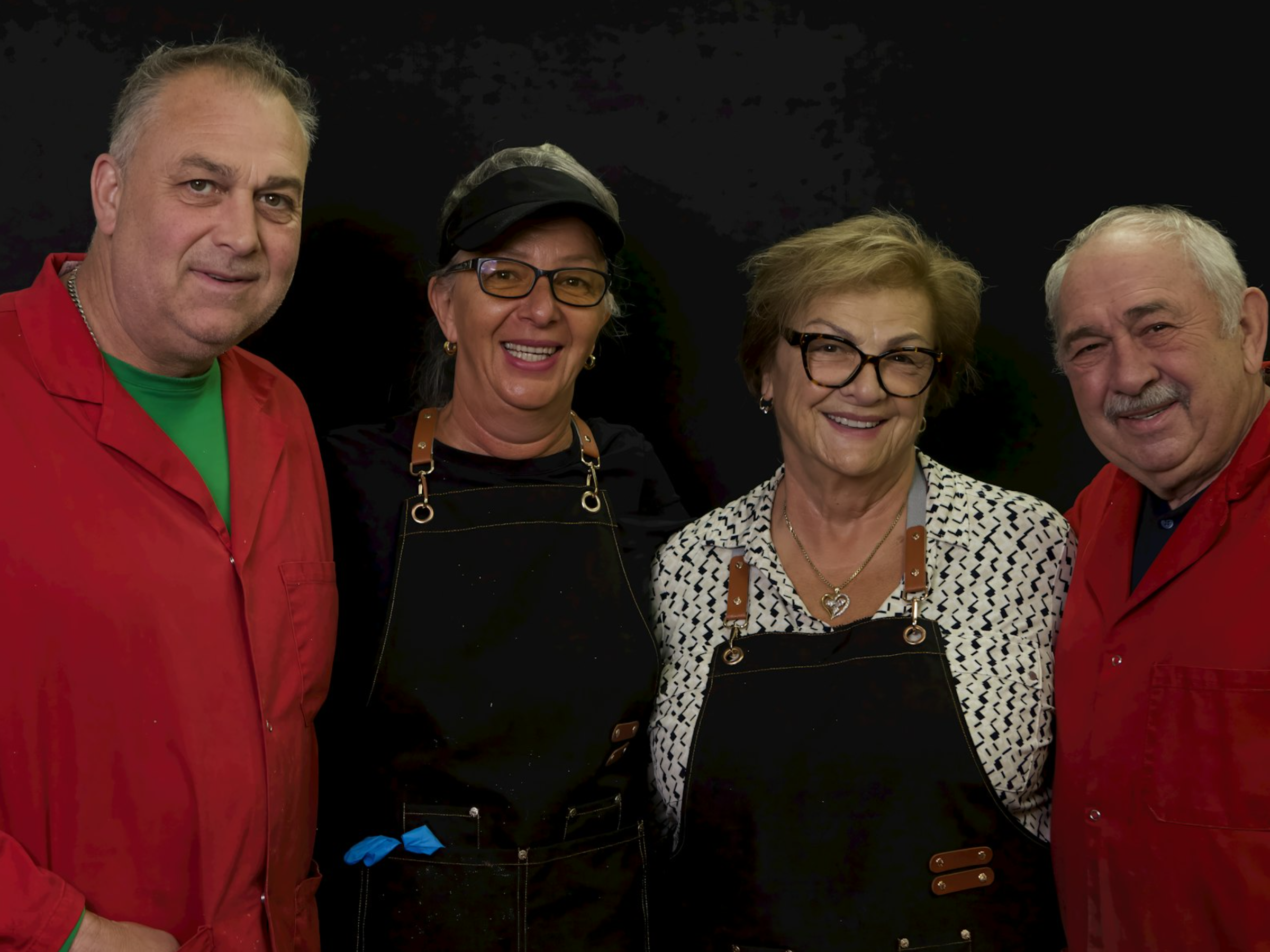 Group of four smiling older adults posing together against a black background, two men and two women, dressed casually, some wearing glasses, and one woman with a chef apron.