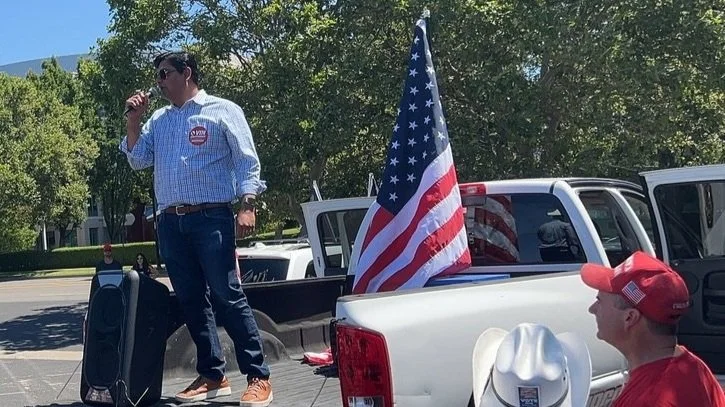 Man speaking into a microphone on a pickup truck with a USA flag, wearing a plaid shirt and campaign badge, outdoor rally setting.