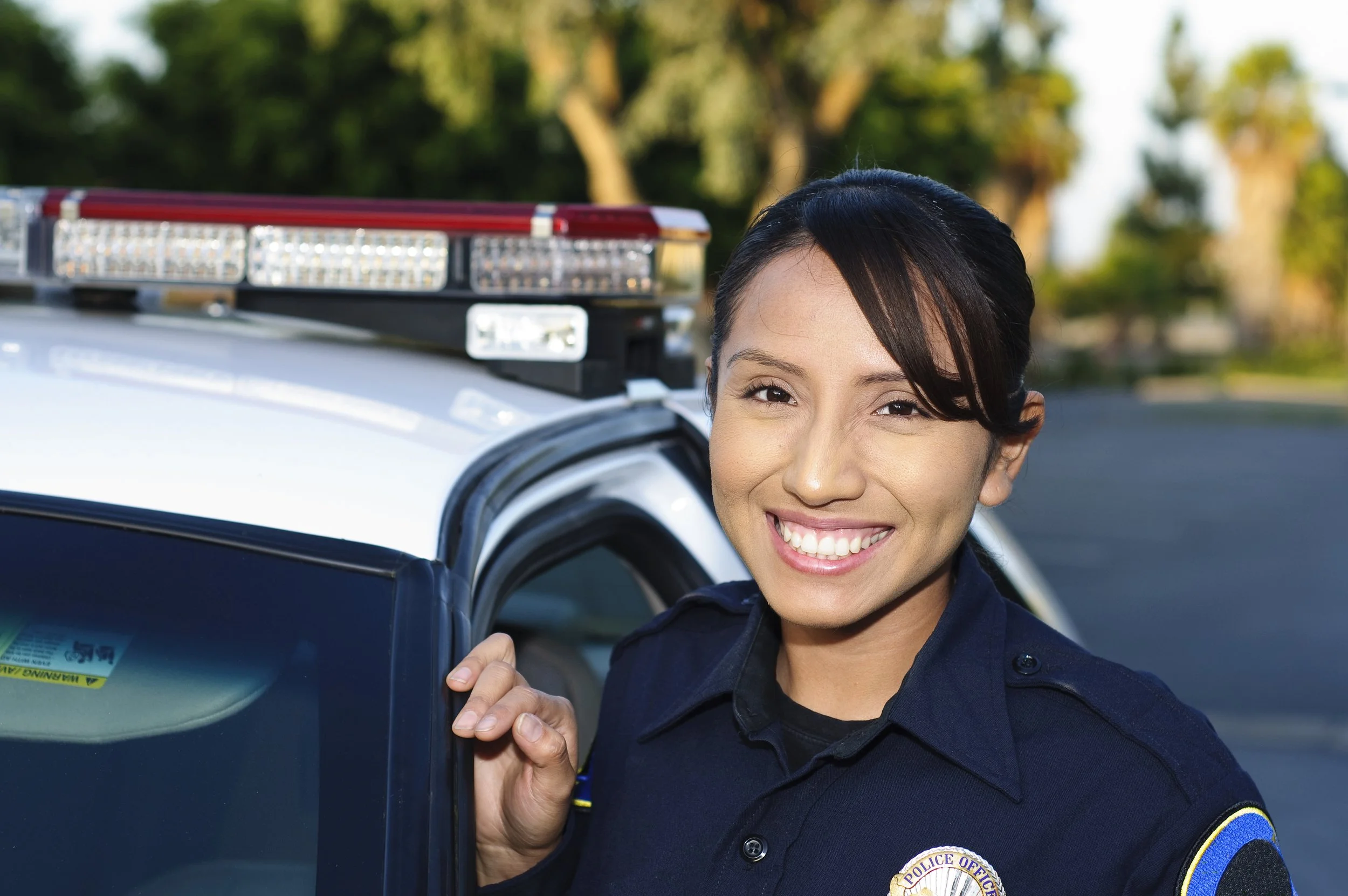 Smiling police officer standing next to patrol car in a parking lot.