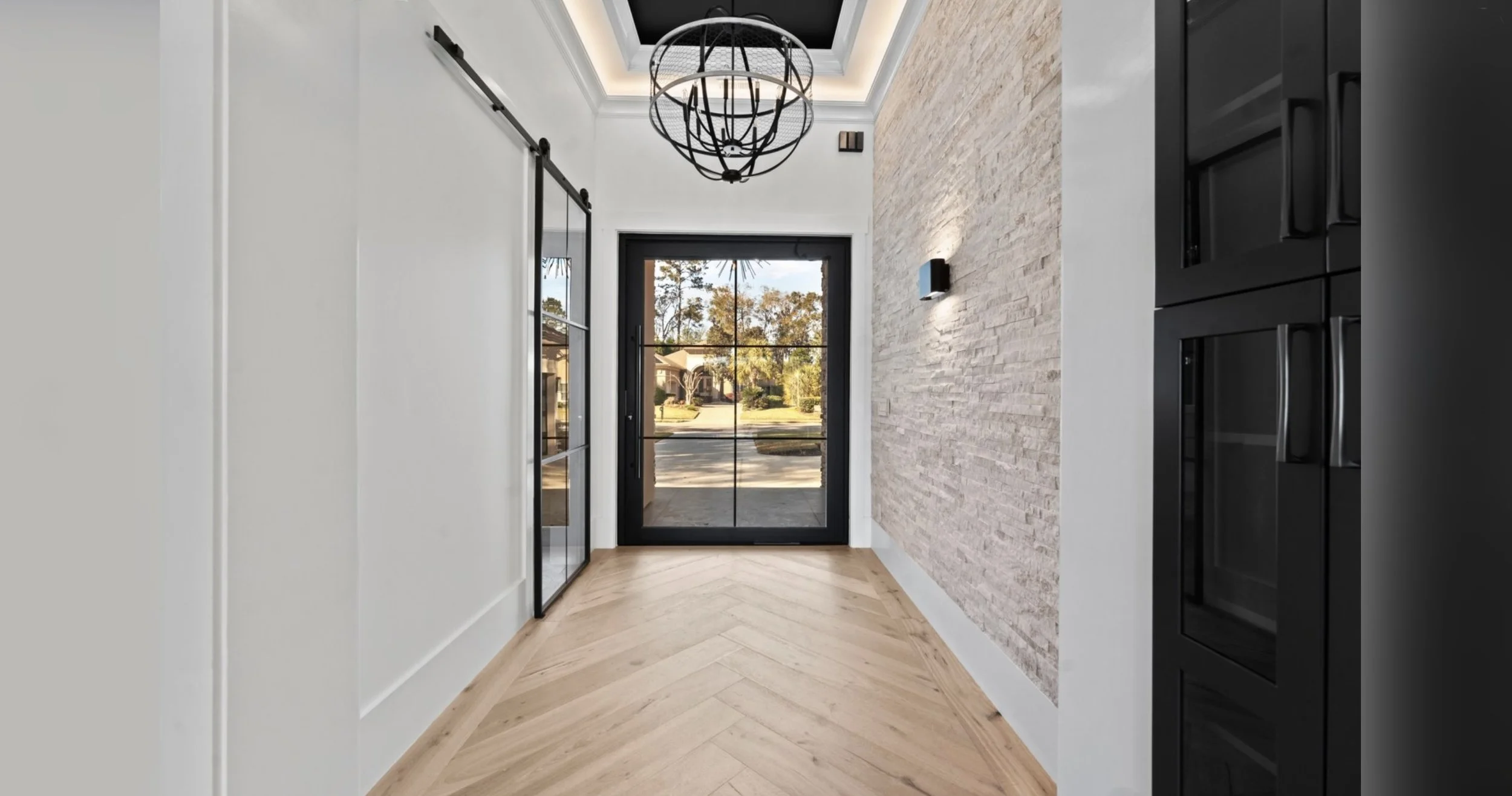 Modern entryway with a glass front door, wood herringbone flooring, a white textured brick wall, black sliding barn door, and a black metal chandelier.