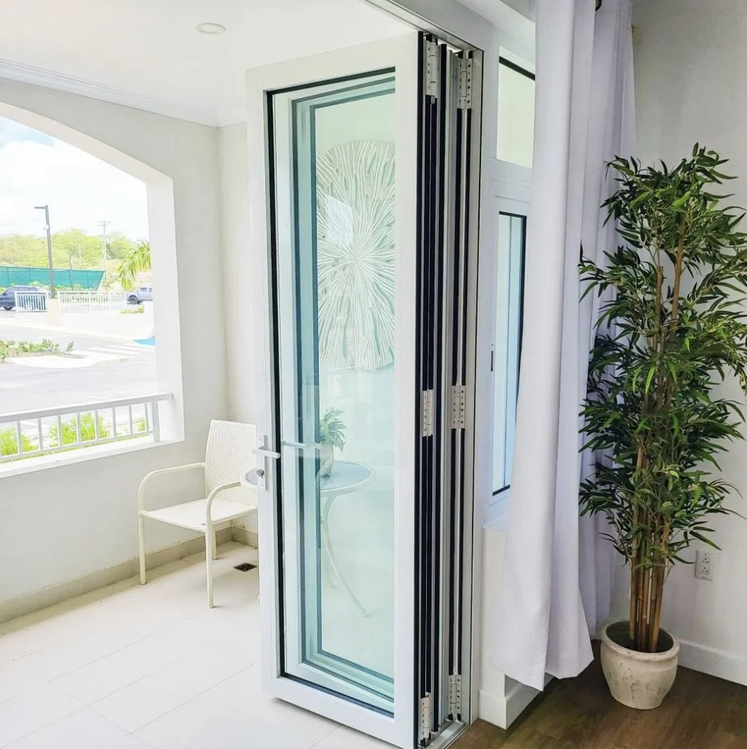 Interior of a room with a large folding glass door, a white chair, a small table with a plant, a window view of a street and greenery, and a tall potted plant in the corner.