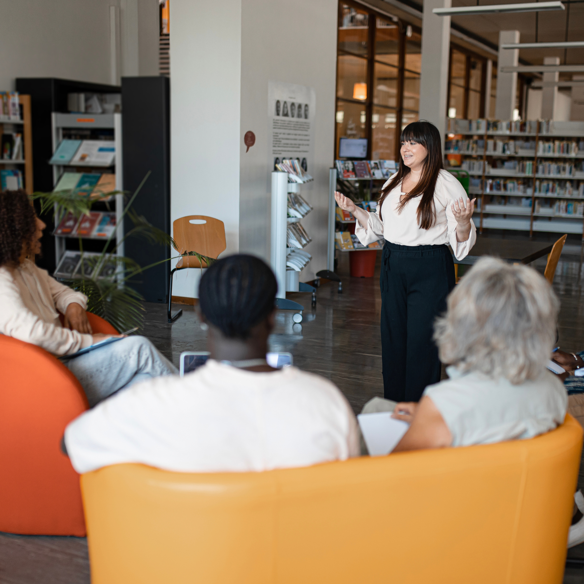 A woman with long dark hair wearing a white blouse and black pants speaking to an audience in a library with bookshelves in the background.
