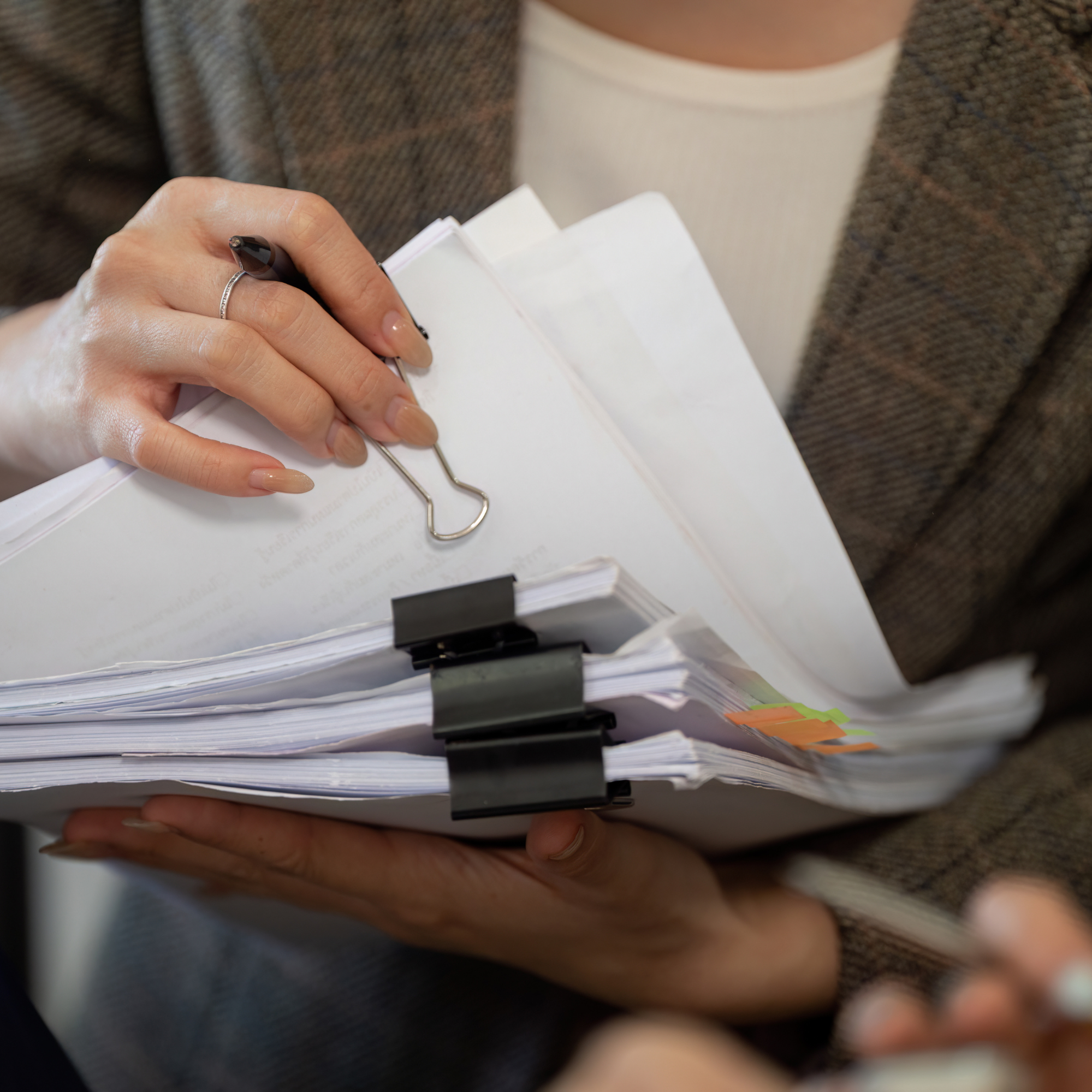 Close-up of a person's hand holding a binder clip on a stack of papers, with a blurred background.