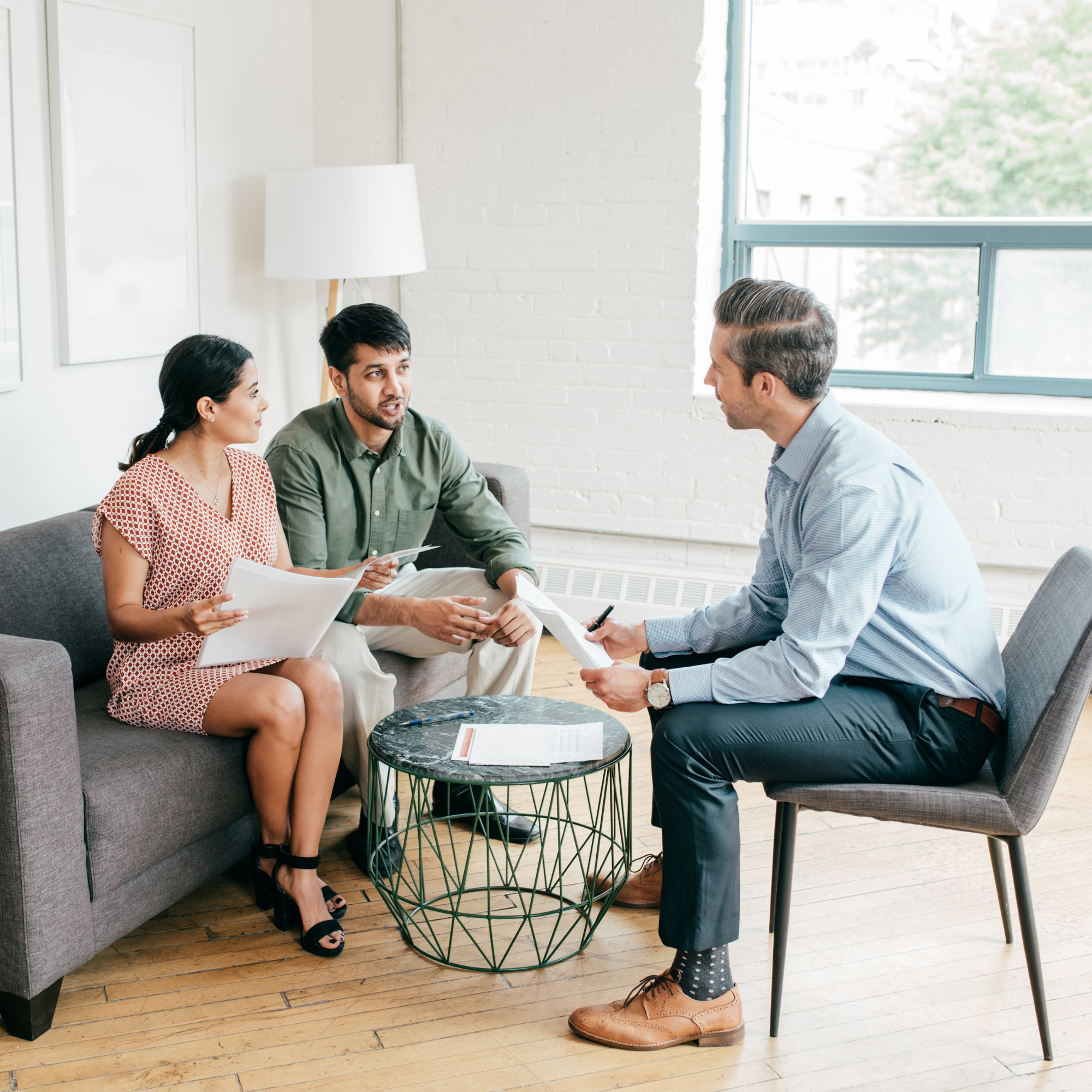 Three people sitting and standing around a small black round table in an office with wooden flooring. They are engaged in conversation, with documents and a pen on the table. A large window with a view of buildings and trees is in the background.