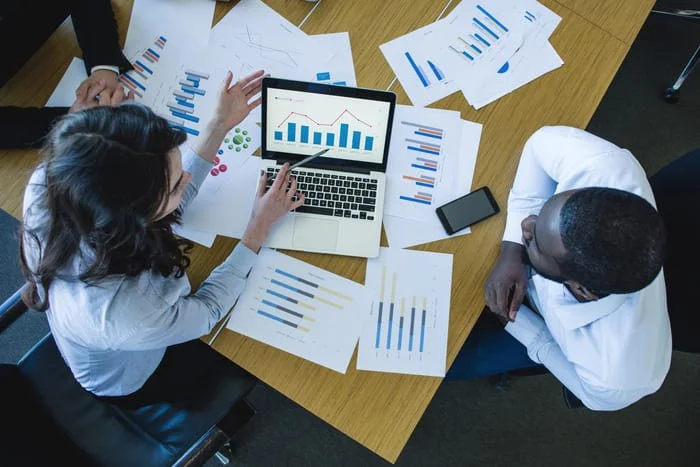Business meeting with two professionals analyzing charts and graphs on paper and a laptop displaying bar and line charts, surrounded by printed reports and documents on a wooden conference table.