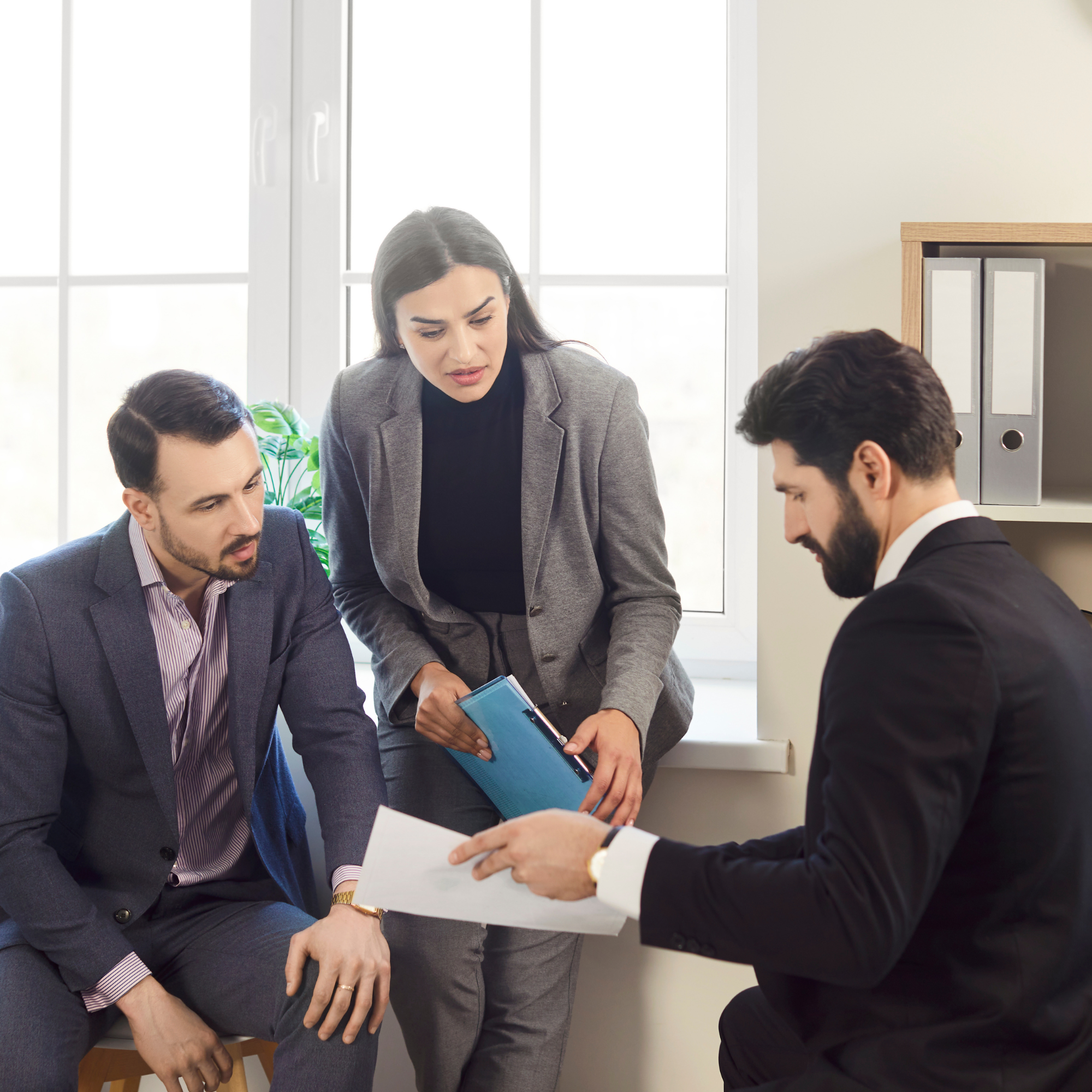 Business professionals in a discussion, with some seated and one woman standing, in an office setting with a window and file organizers in the background.