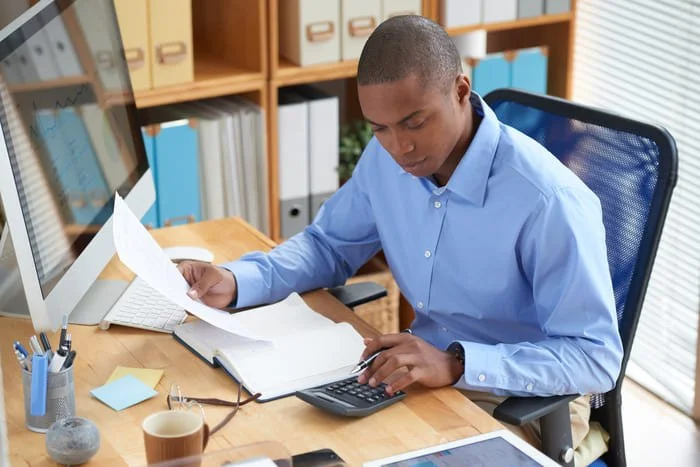 A man in a blue shirt working at a desk with a computer, papers, and office supplies in a home office.
