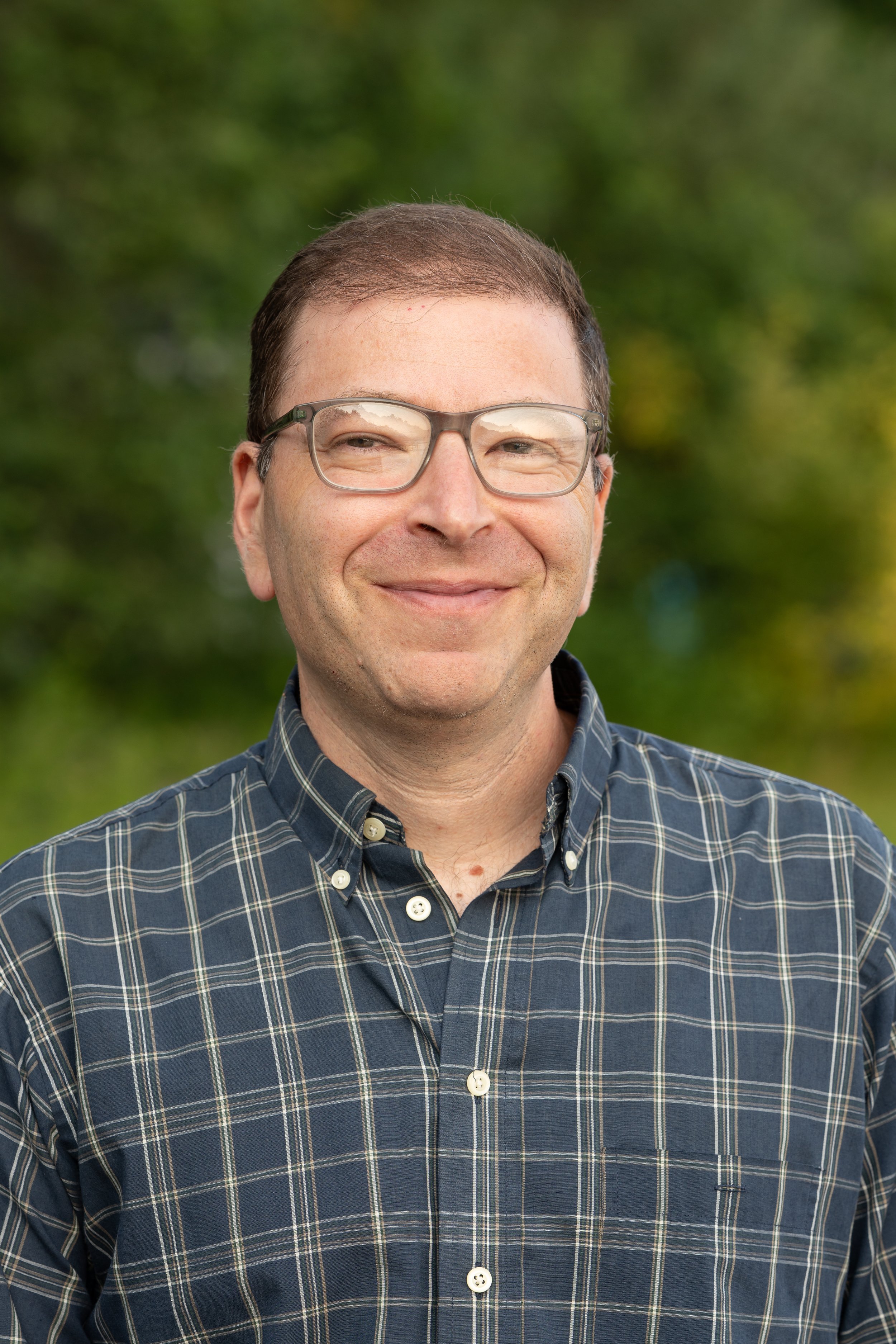 Headshot of a man smiling outdoors wearing glasses and a blue plaid shirt.