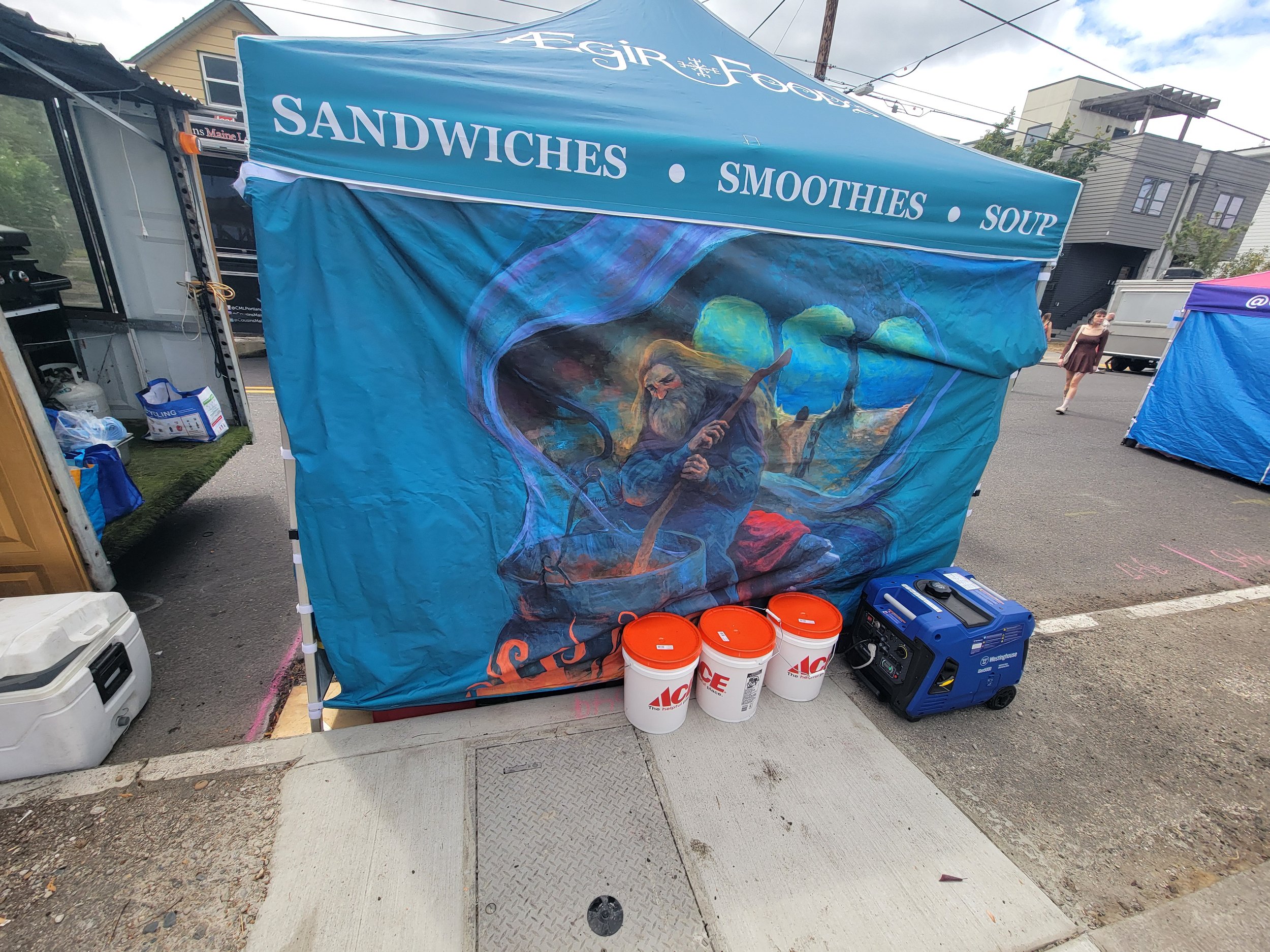 A street-side food stall with a blue canopy displaying the words 'SANDWICHES,' 'SMOOTHIES,' and 'SOUP.' The stall features a colorful illustrated banner of a wizard or sorcerer stirring a cauldron. In front are three white buckets labeled 'ACE,' a blue toolbox, and boxes of supplies, with a woman walking in the background.