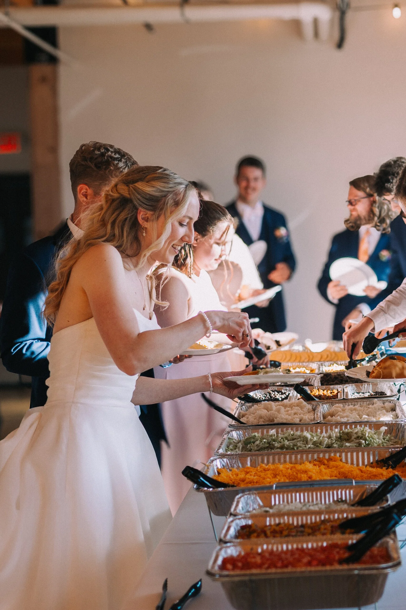 People in formal attire, including a woman in a wedding dress, serve themselves food from buffet trays at a wedding reception.