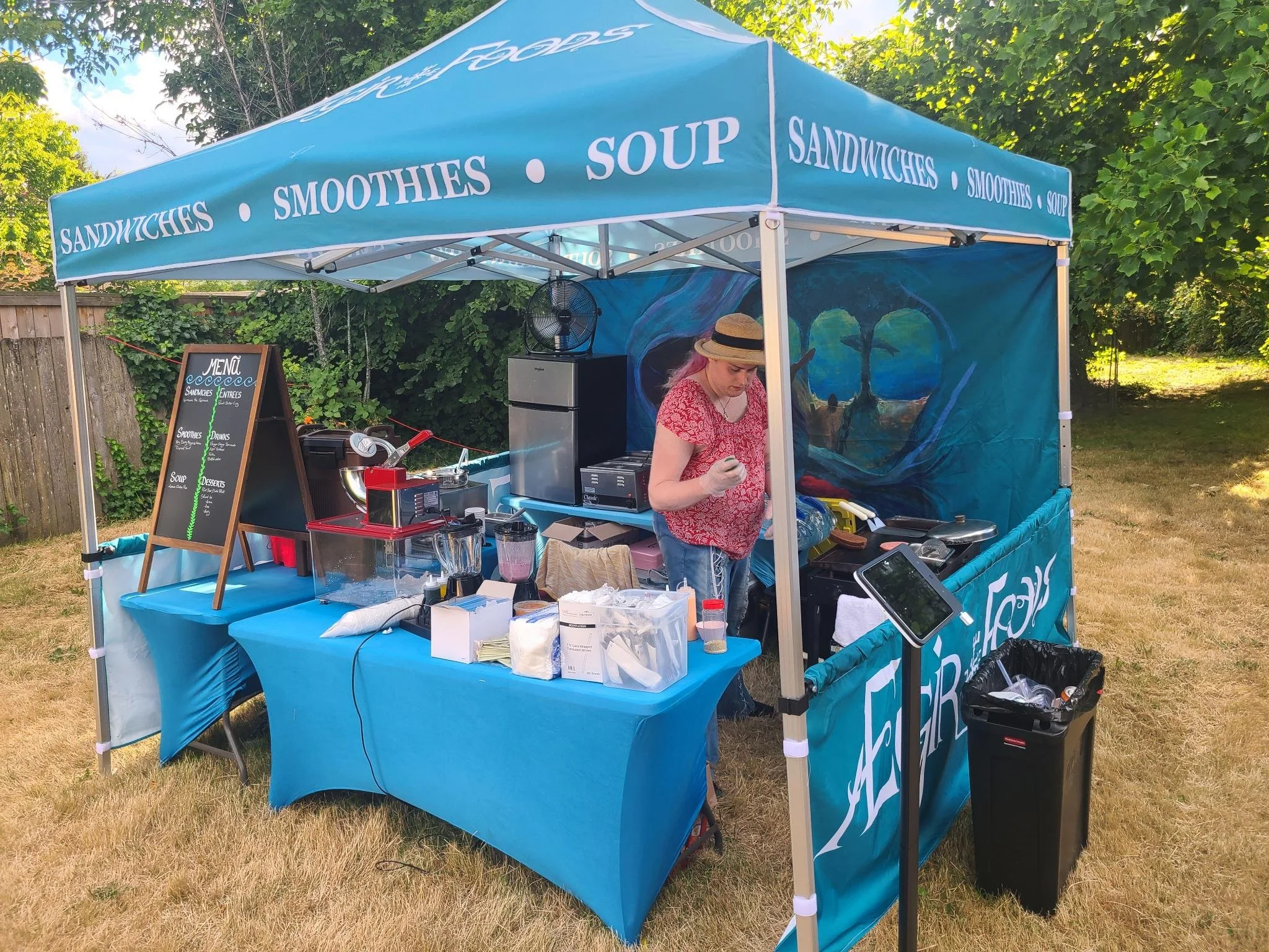 A person preparing food at a blue outdoor food stand selling sandwiches, smoothies, and soup, with a menu board, surrounded by trees and grass.