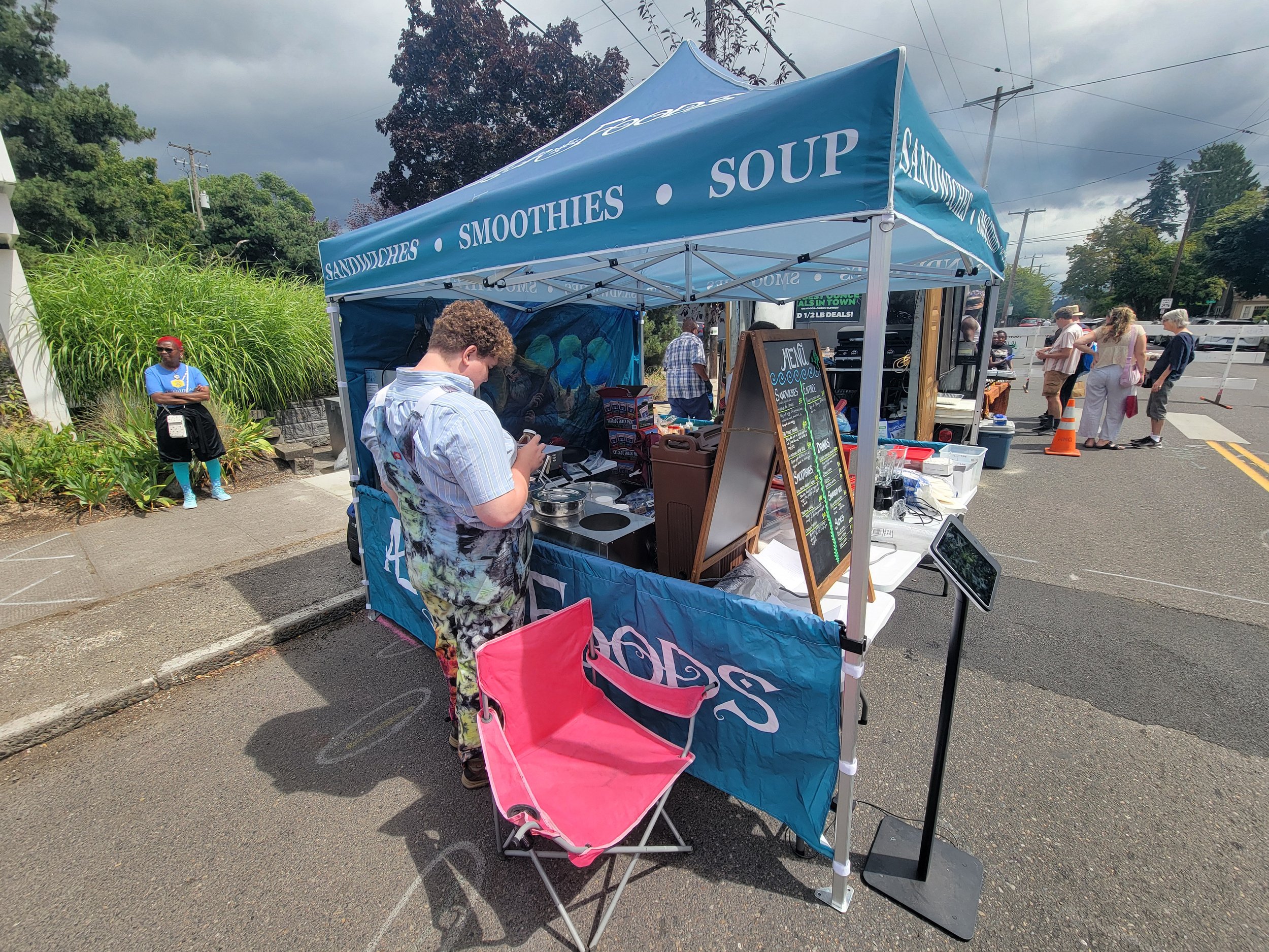 Outdoor stand with blue canopy selling smoothies, sandwiches, soup, and sandwiches with a menu board, with people browsing and a pink chair in front.