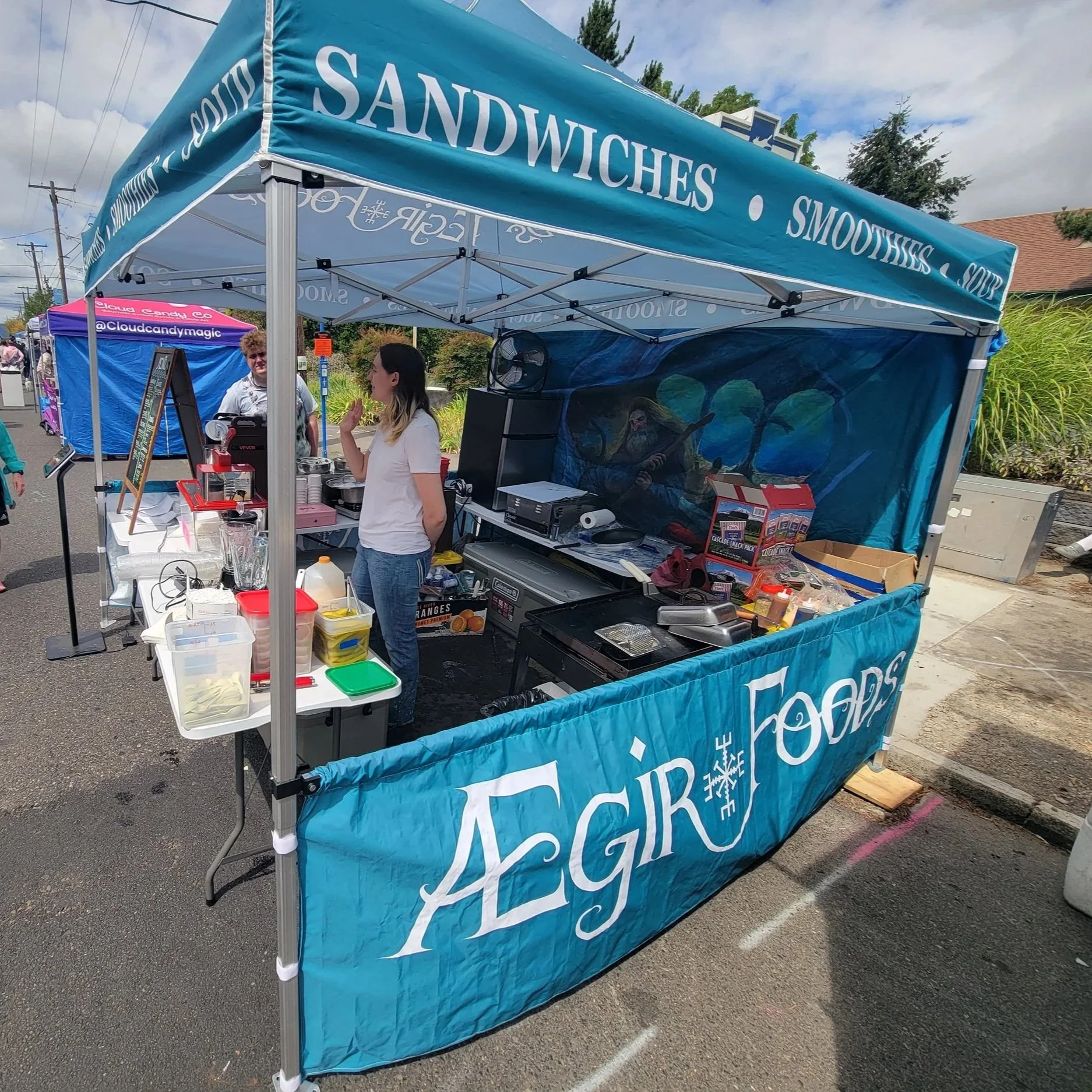 A blue outdoor booth canopy with the words 'SANDWICHES,' 'SMOOTHIES,' and 'SOUP' printed on it. Behind the booth, a woman is talking to a person standing nearby. The booth has various supplies and equipment for serving food and beverages, and an artistic mural of a wizard or mage playing a guitar painted on the back wall of the booth.
