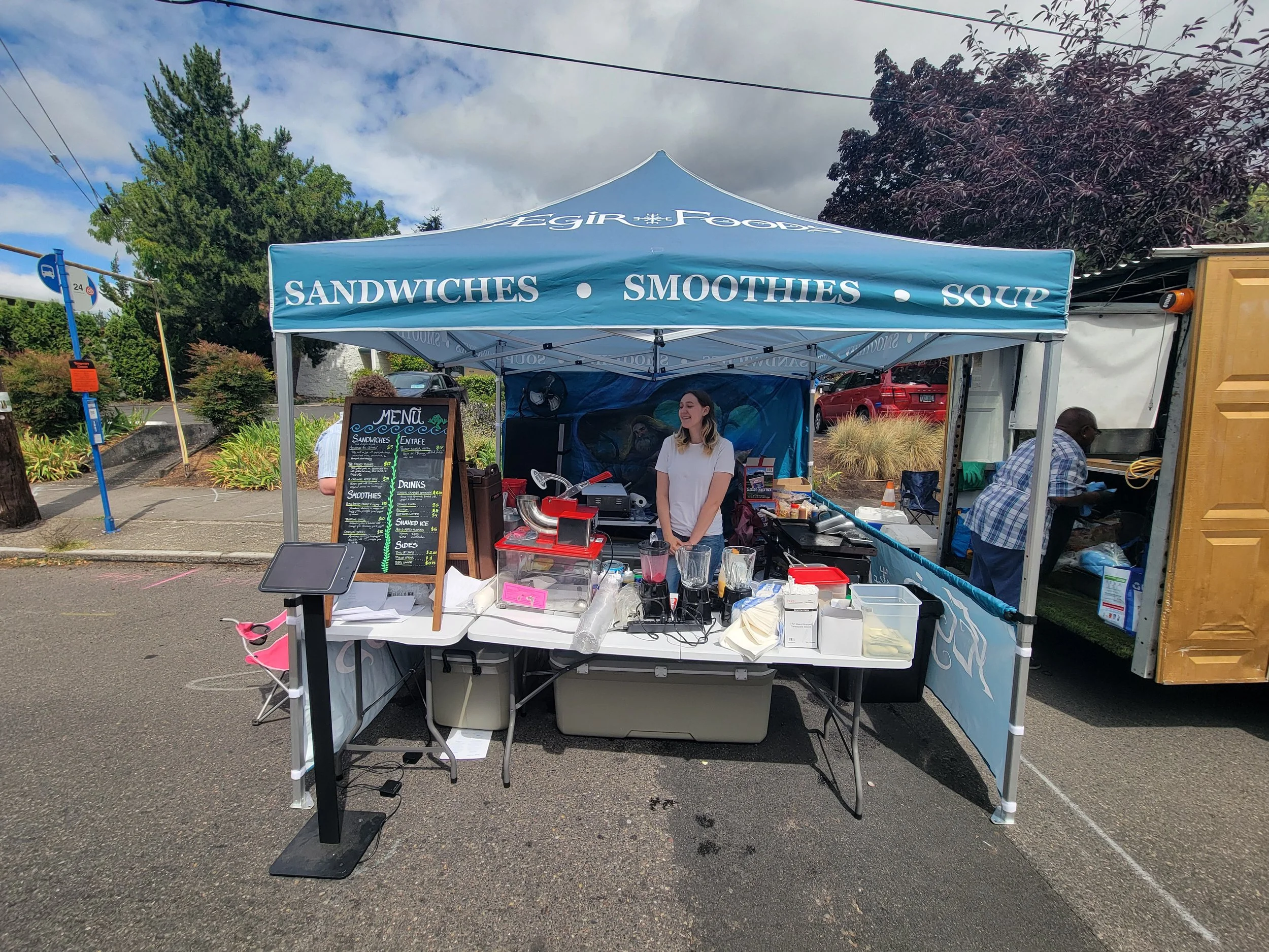 A food stand with a blue canopy labeled 'Sandwiches, Smoothies, Soup' at an outdoor market, with people and trees in the background.