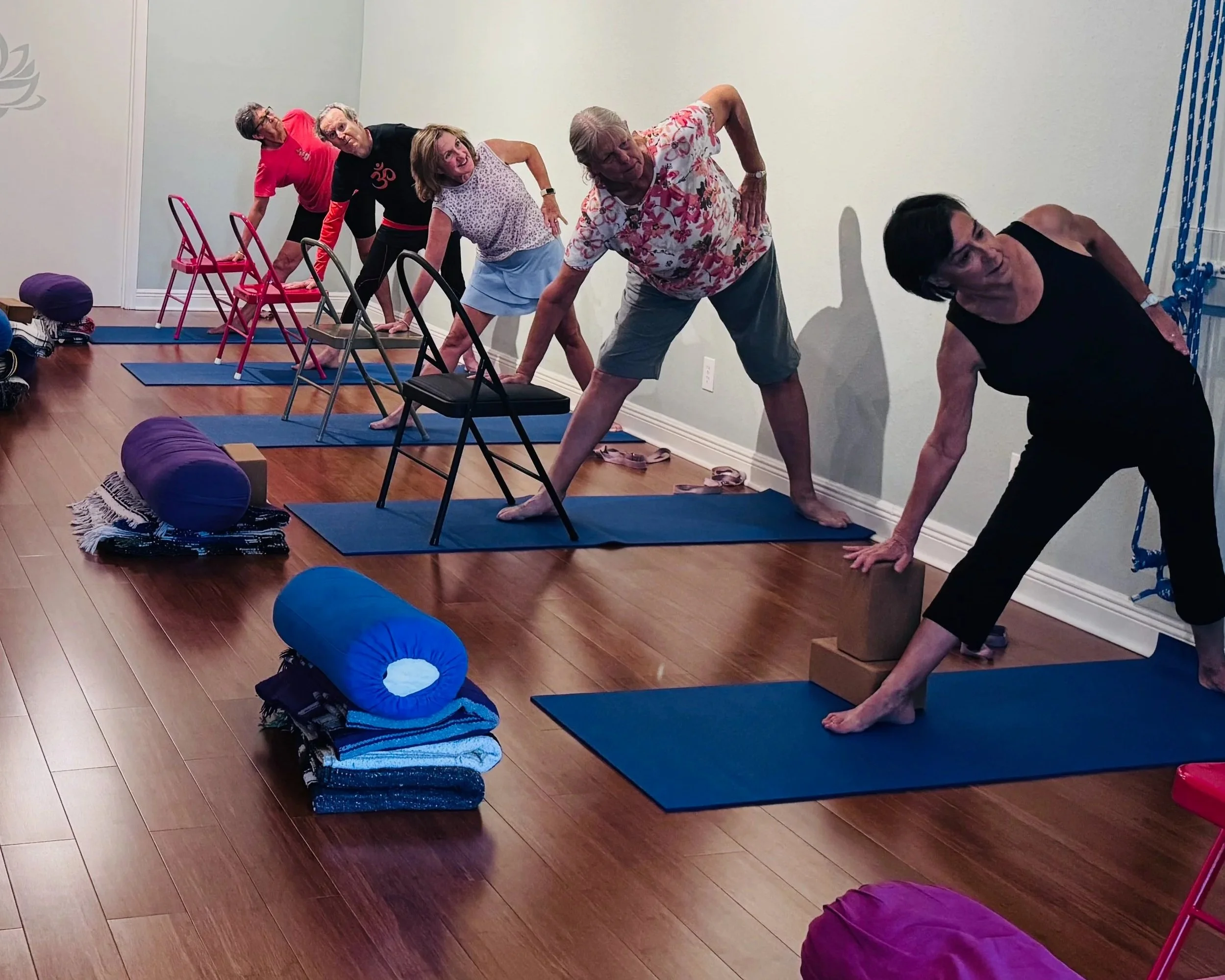 Elderly people doing yoga using chairs for support in a side stretch pose.