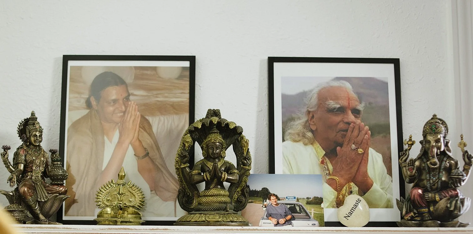 A shrine with two large framed photographs of elderly Indian spiritual leaders, decorated with Hindu statues, a photo of a man next to a car, and a stone with the word 'Namaste' written on it.