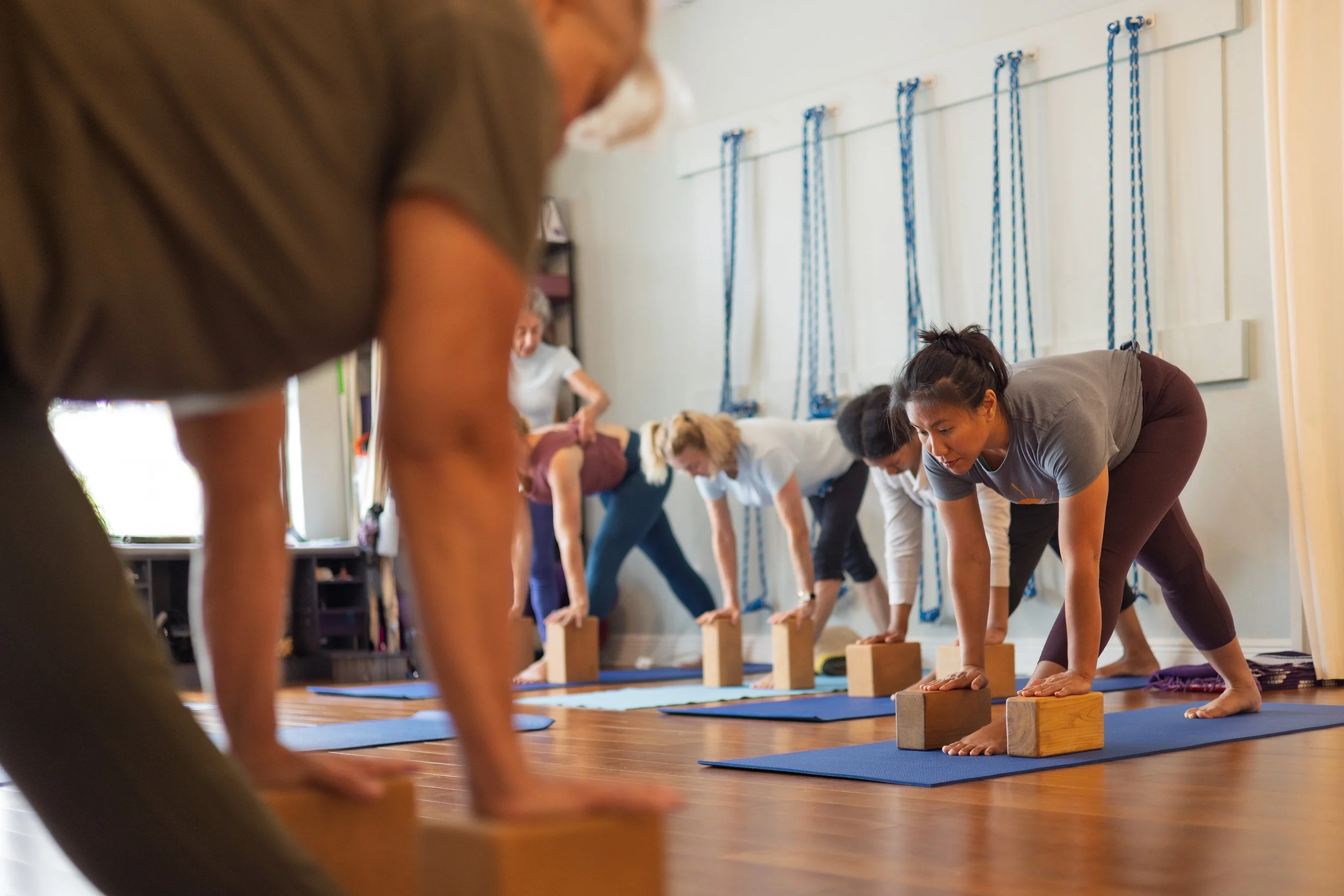 People participating in a yoga class, performing forward bends on mats with wooden blocks as support in a well-lit room with wooden floors and a wall with blue ropes.
