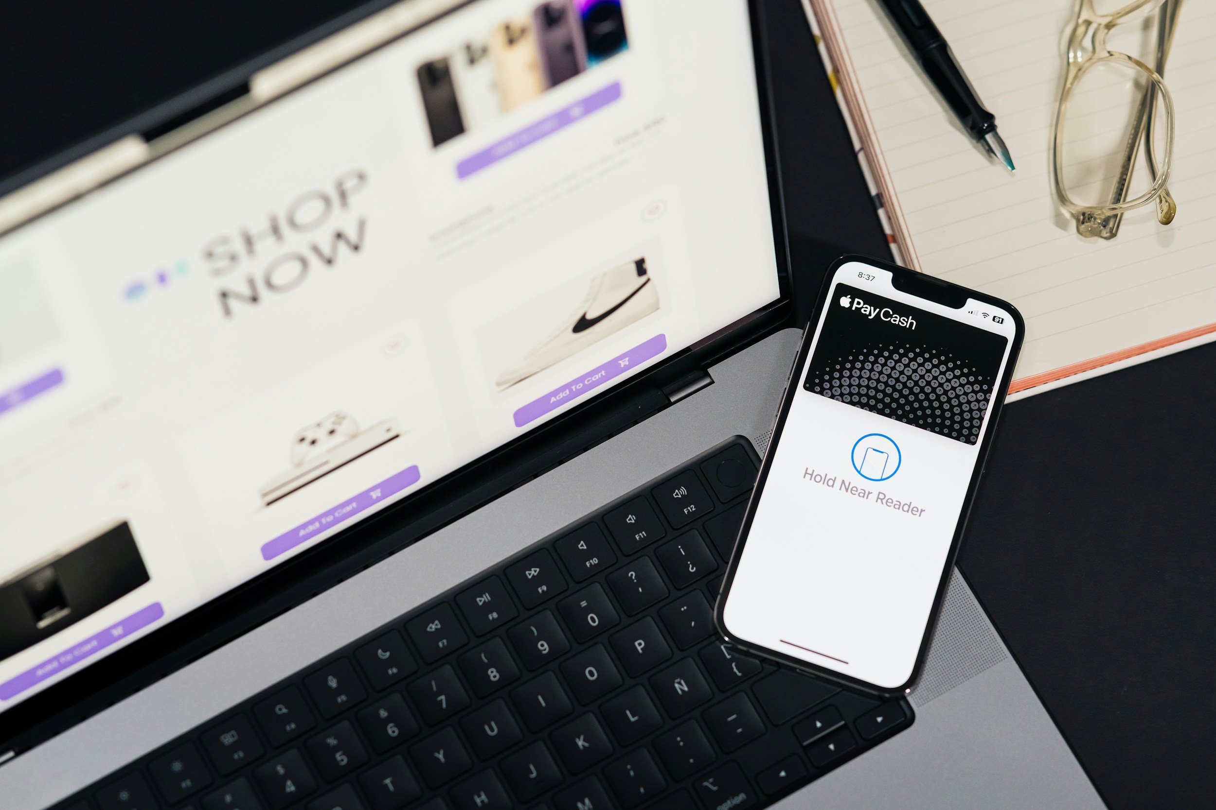 A laptop computer displaying an online shopping website with the words 'SHOP NOW'. A smartphone showing a mobile payment app with a notification to hold near the reader. Next to the devices, there is a notebook with a pen and a pair of eyeglasses placed on it.