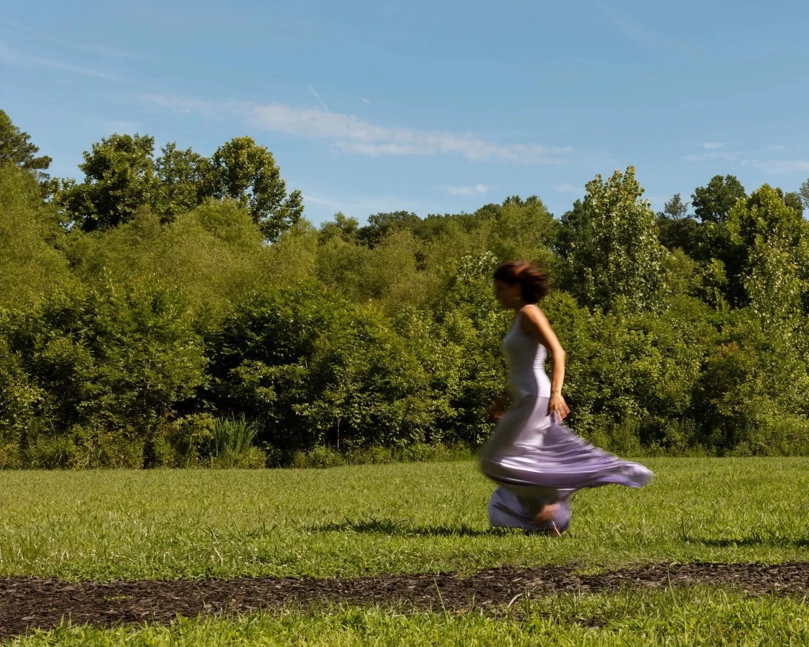 A woman in a long white dress running across a grassy field with a backdrop of green trees and a blue sky.