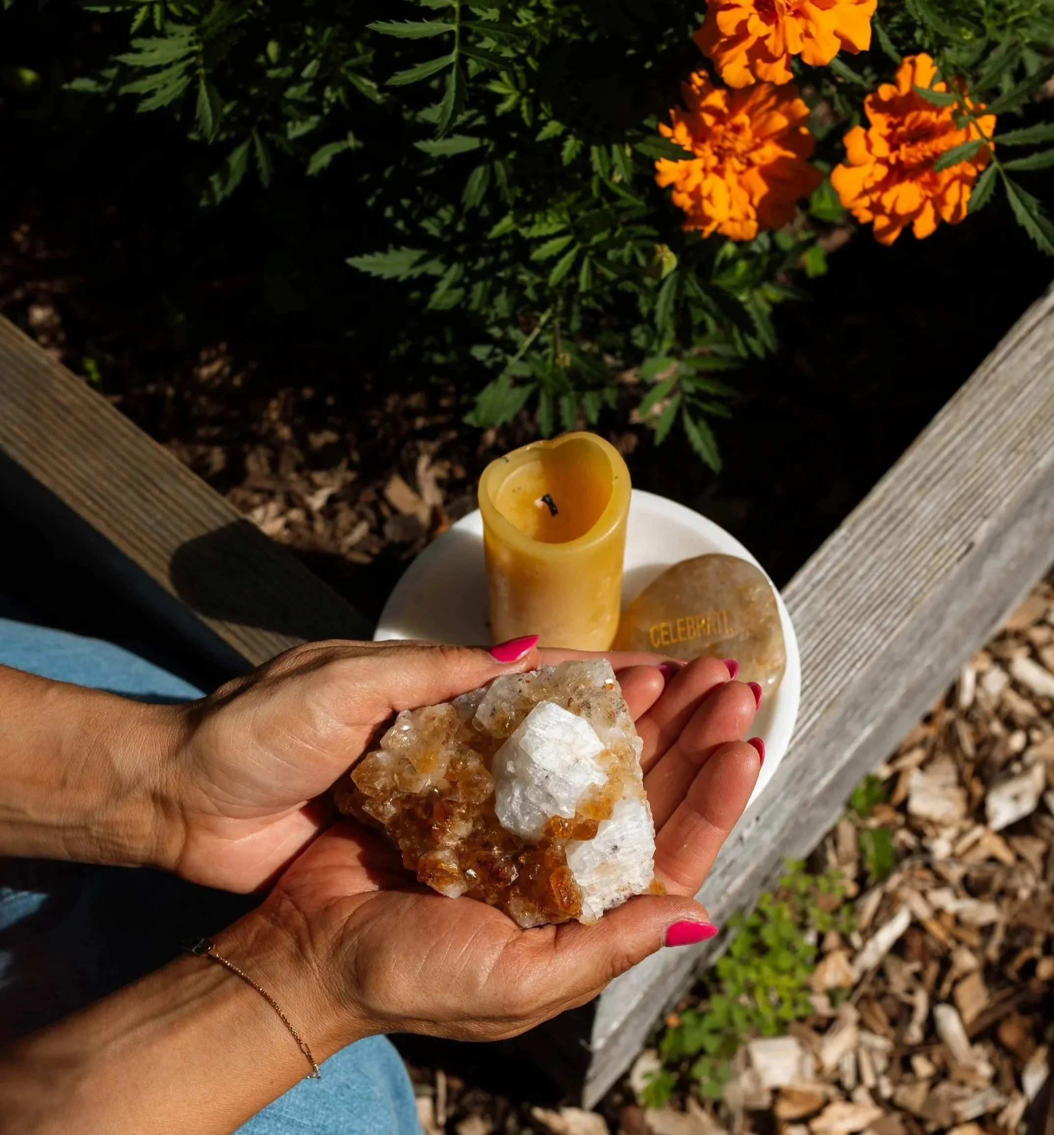 A person's hand holding a large crystal cluster with brown and white minerals, a yellow candle, and a white base with the word 'CELEBRATE' engraved, against a background of orange marigold flowers and green foliage.