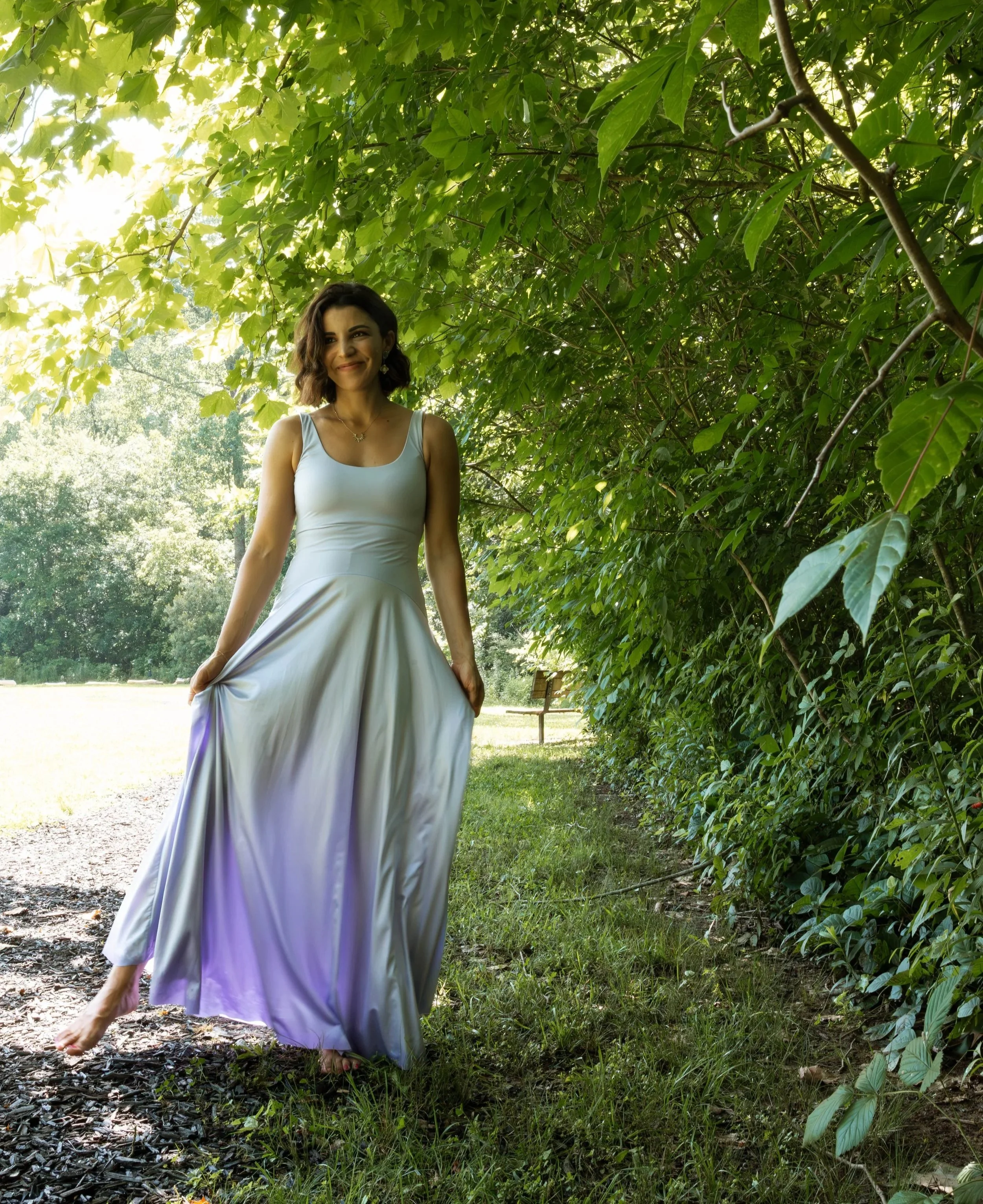 A woman in a white dress holding the bottom of her dress with one hand walking on a grassy path in a lush green park or garden surrounded by dense leafy trees.