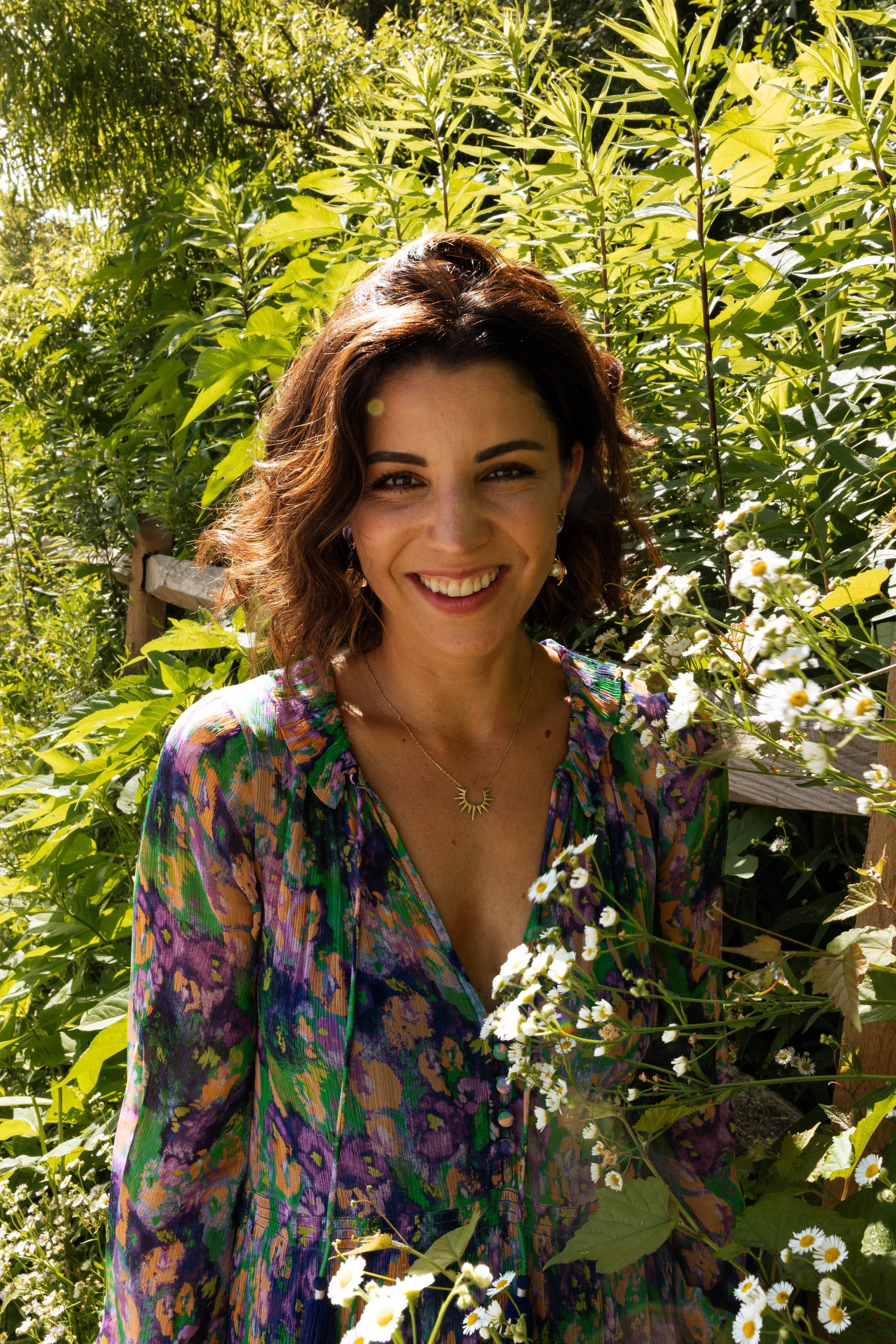 A woman with brown, curly hair, smiling, standing among green plants and white daisies, wearing a colorful, patterned blouse and a moon-shaped necklace, in outdoor sunlight.