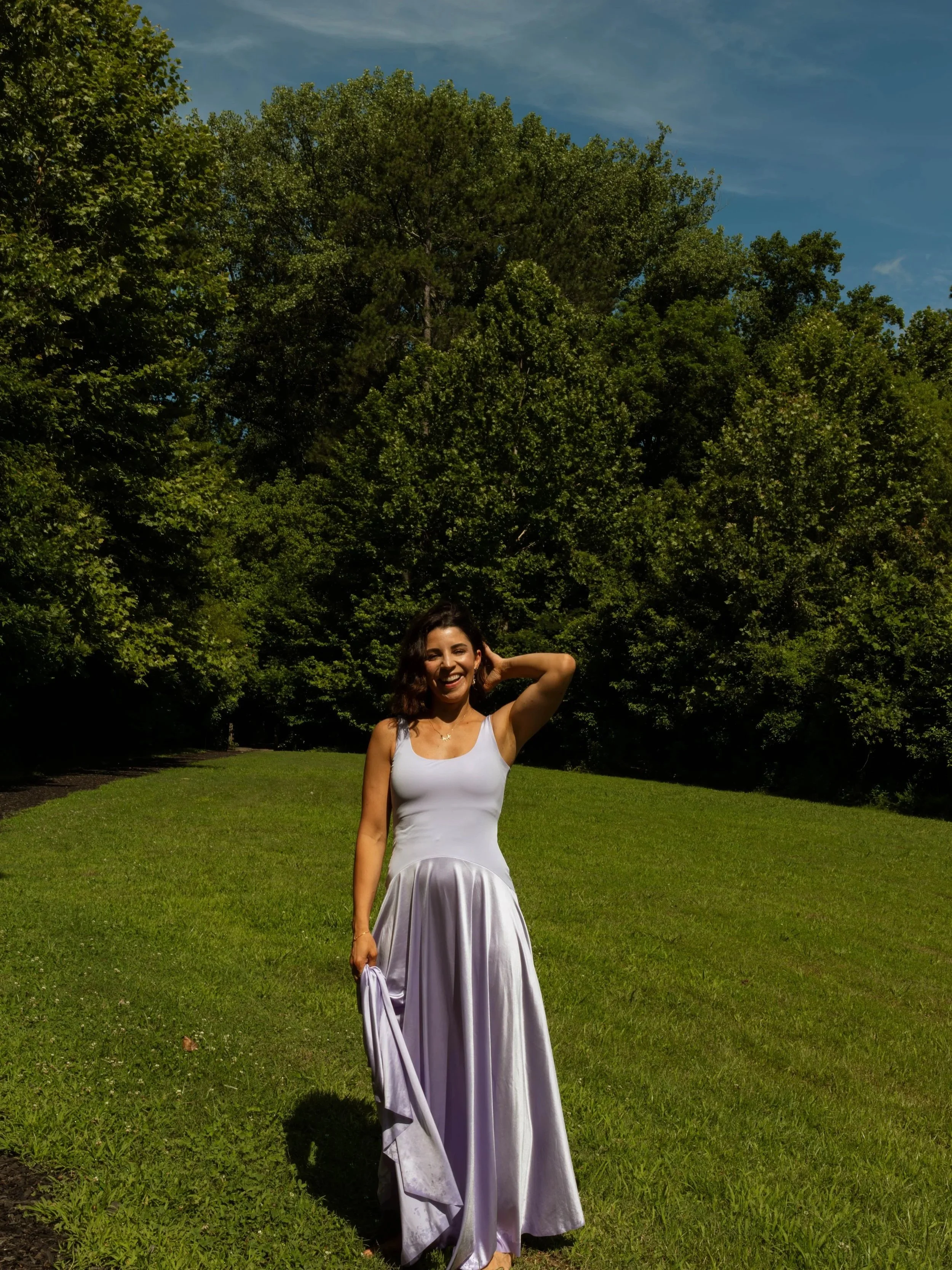 A woman in a white dress standing on a grassy field with trees behind her and a blue sky above, smiling and posing with one hand near her head.