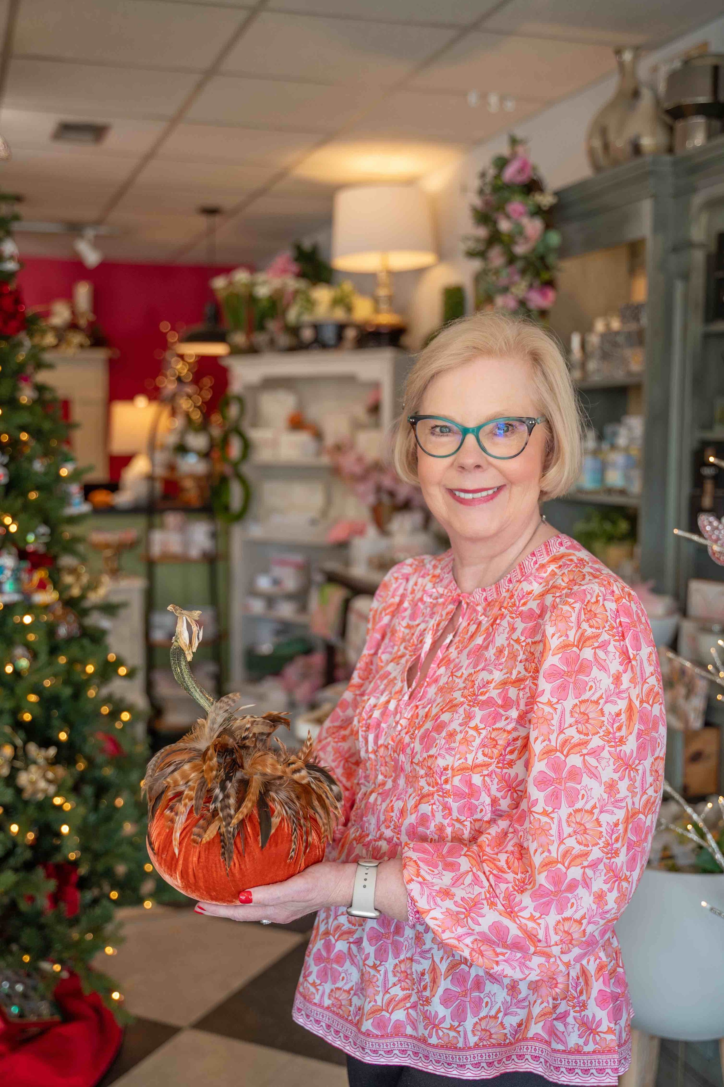 A smiling woman wearing glasses and a pink floral dress, holding a pumpkin decorated with feathers, in a room with Christmas decorations and a Christmas tree.