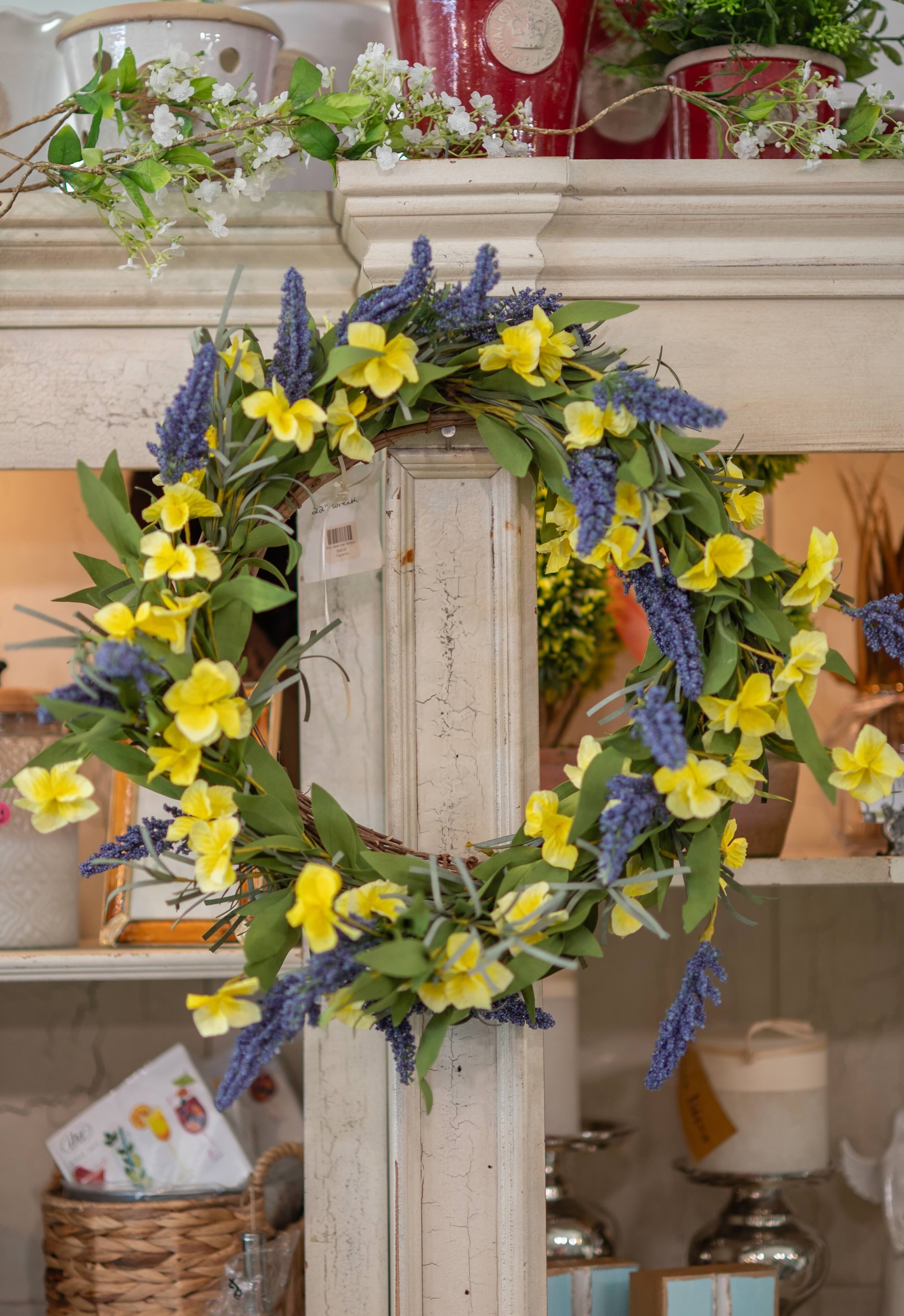 A decorative white wooden frame with a floral wreath made of yellow and purple flowers hanging on it. In the background, there are various home decor items including potted plants, glass jars, and baskets.