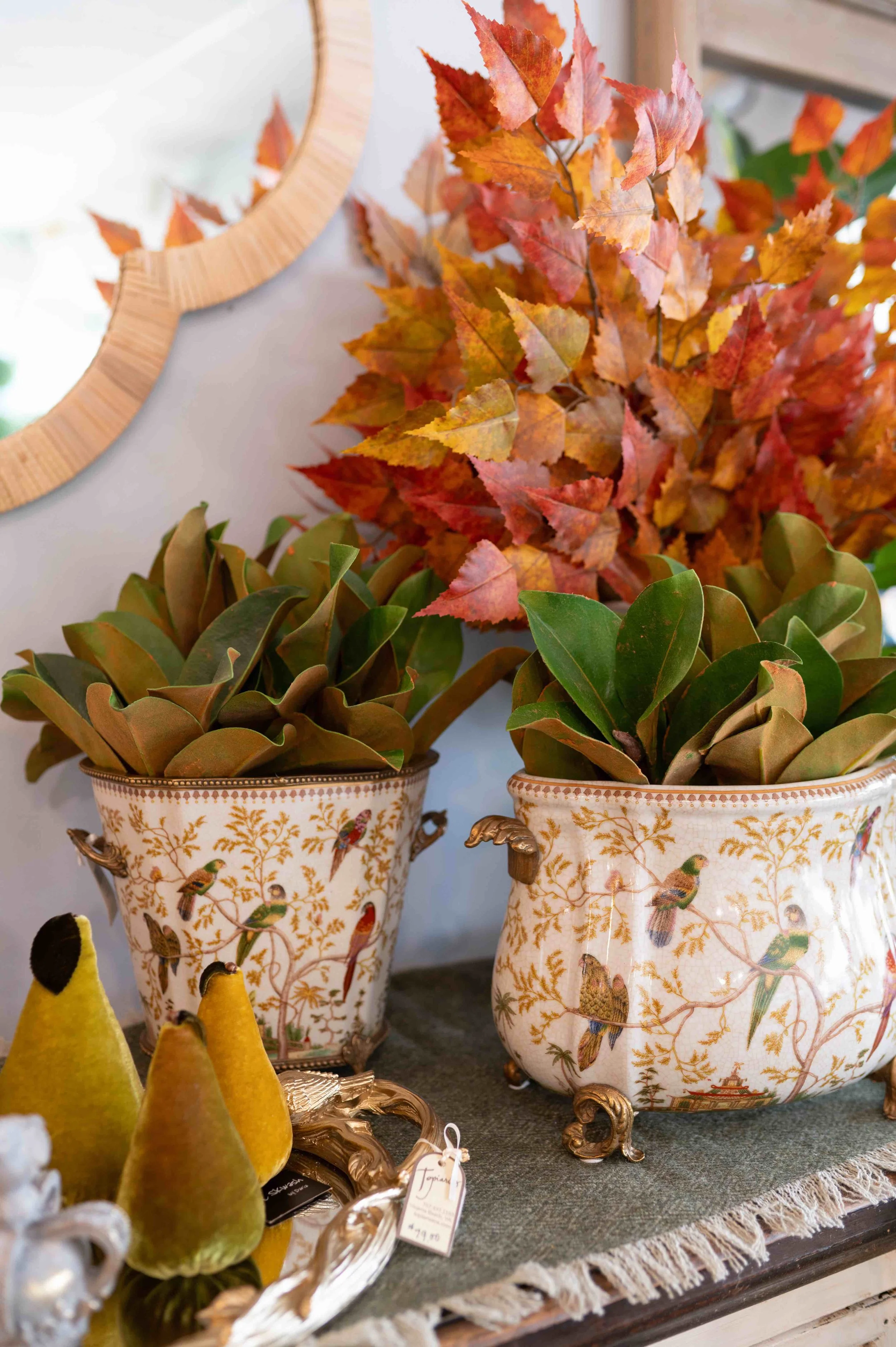Decorative porcelain vases with bird patterns, filled with artificial autumn leaves and surrounded by yellow felt peapods, displayed on a woven cloth surface.