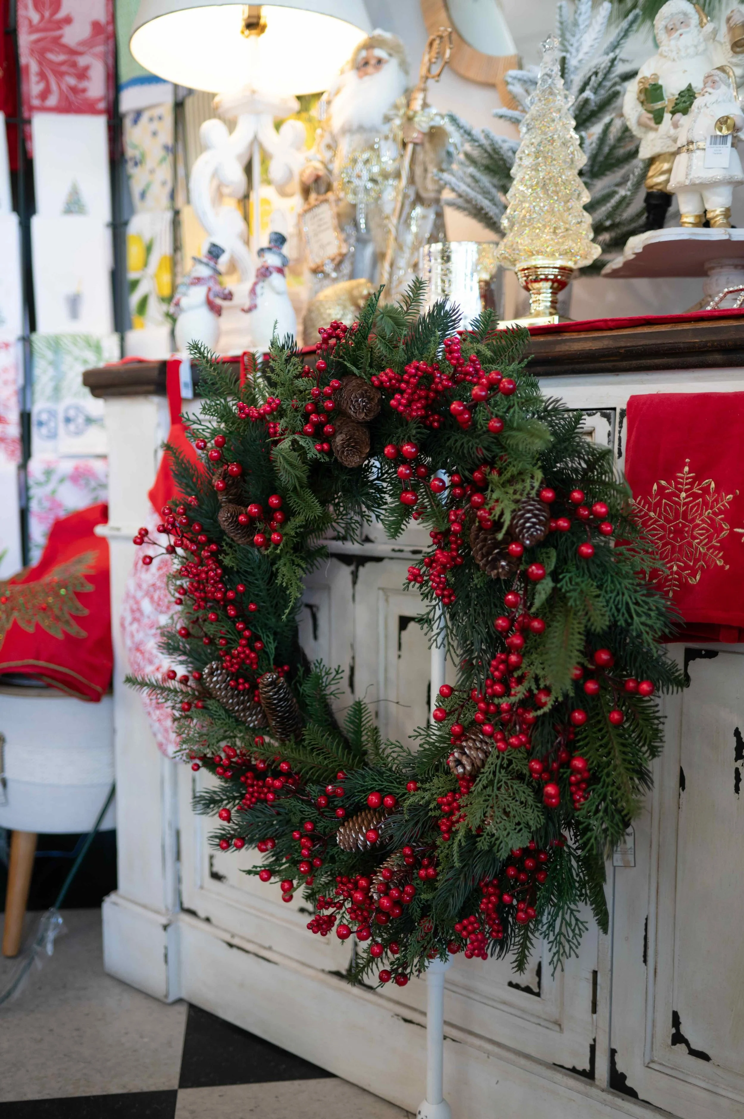 A Christmas wreath made of evergreen branches, red berries, and pinecones hanging in front of a white cabinet. The background features various Christmas decorations, including Santa figurines, a gold glittery Christmas tree, snowmen, and holiday items.