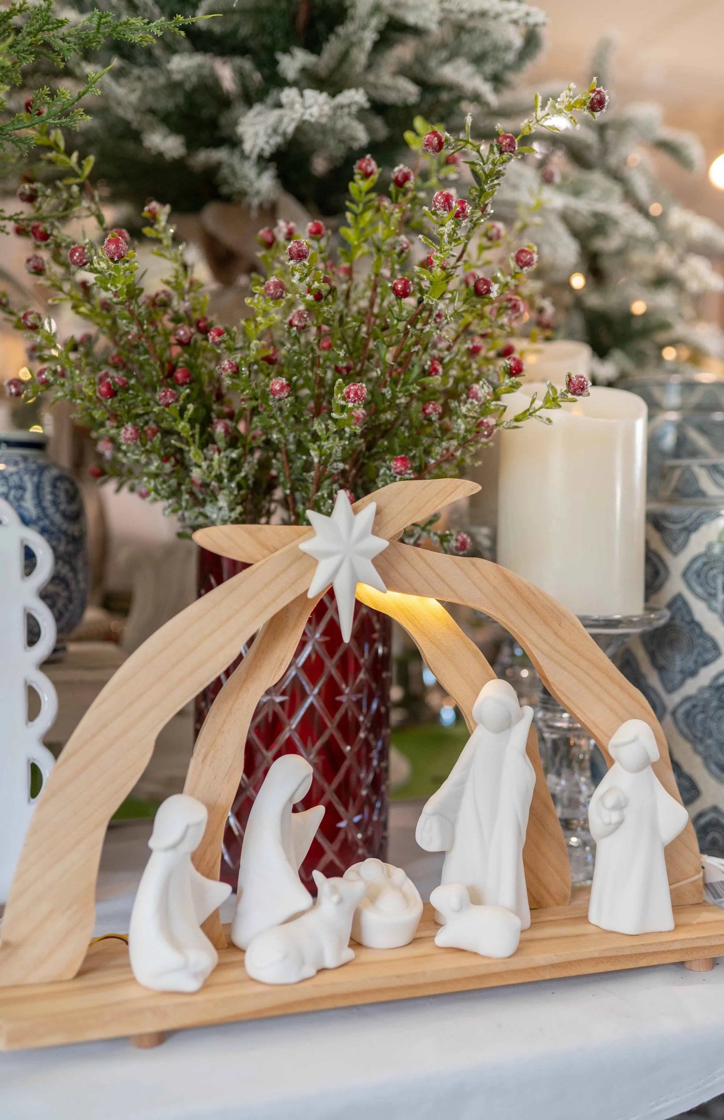Nativity scene with white figurines under a wooden arch, with Christmas decorations and candles in the background.