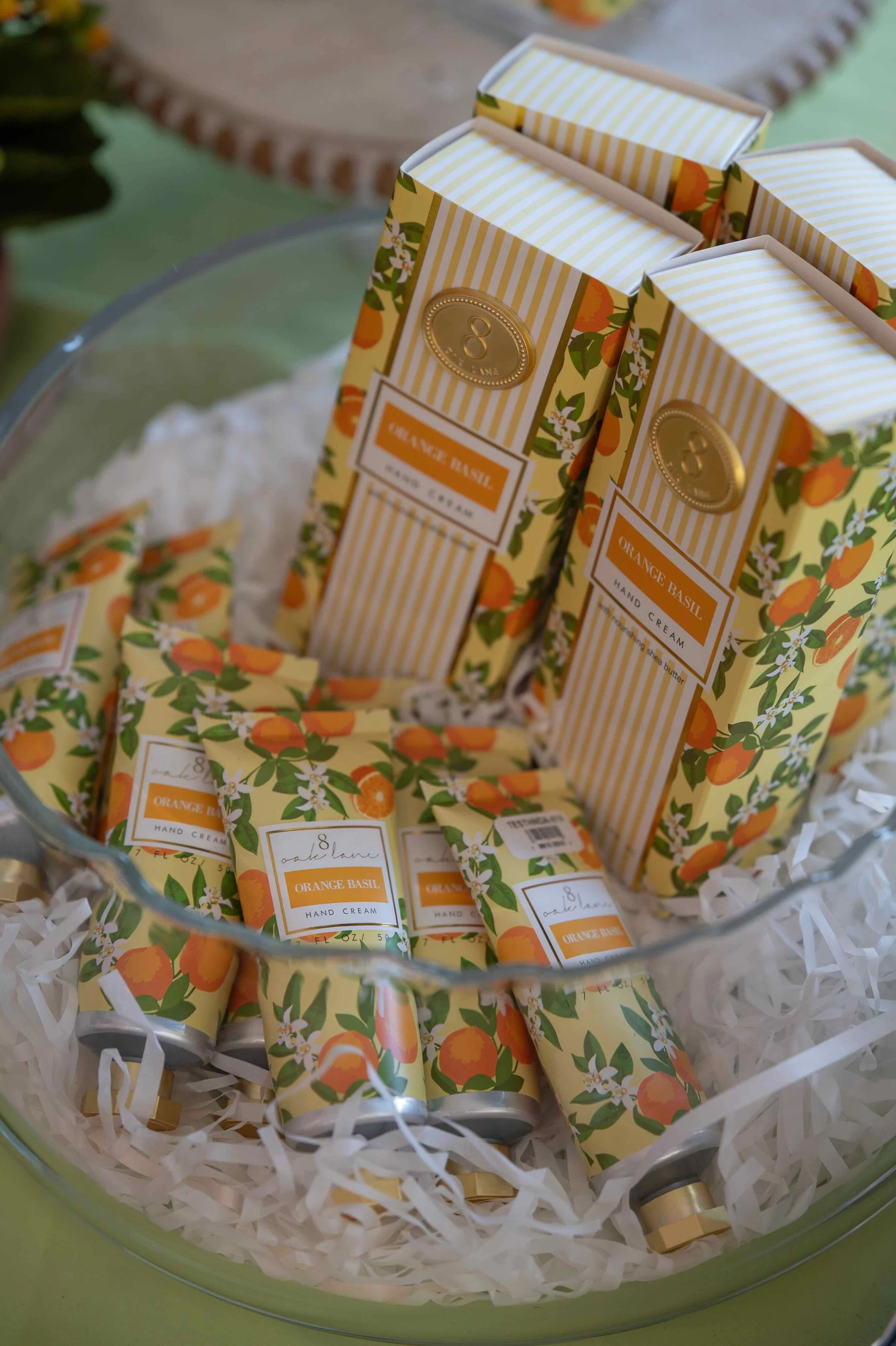 Multiple bottles of Orange Basil hand cream in floral packaging arranged in a glass bowl with shredded paper inside.