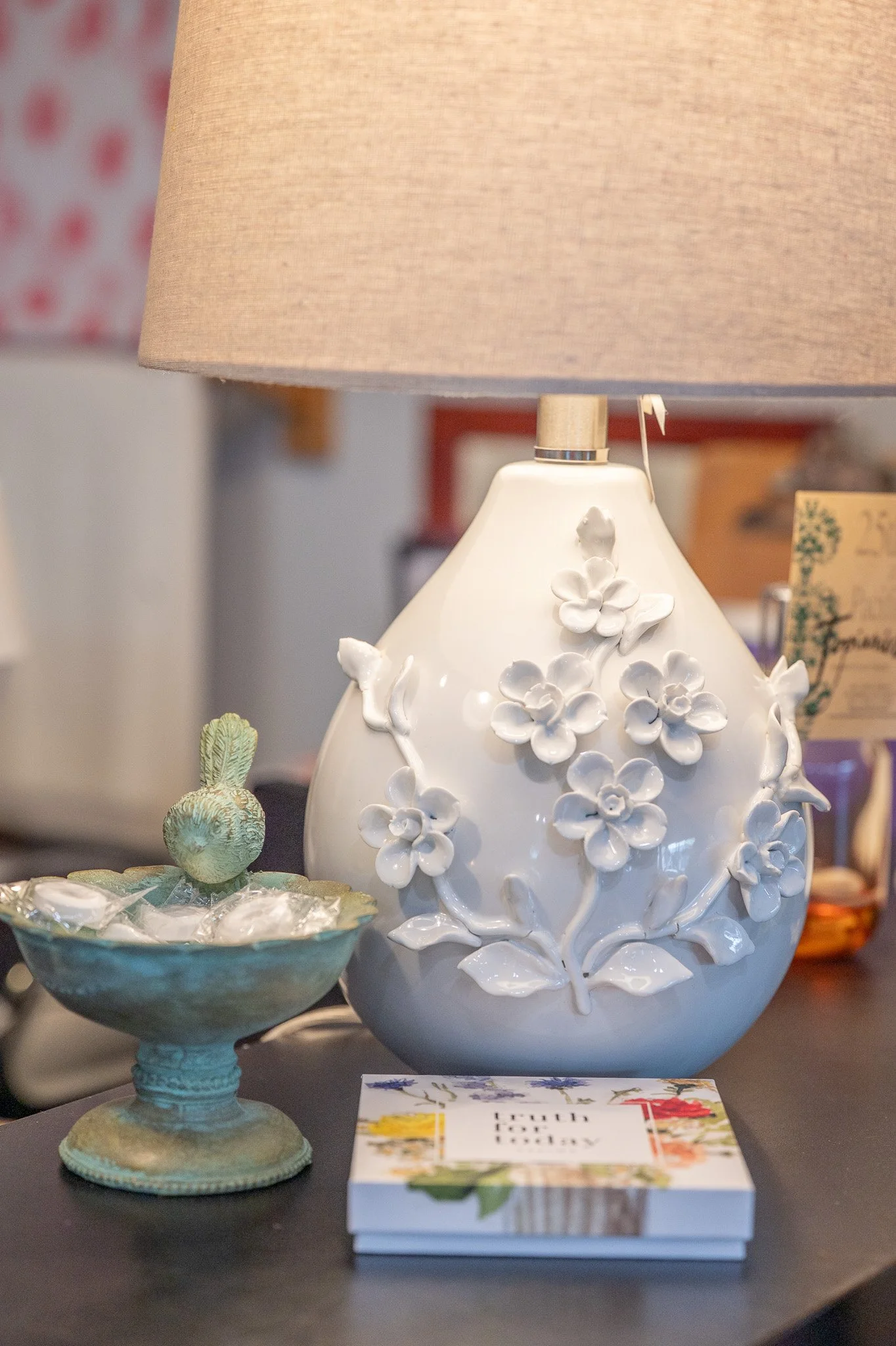 A white ceramic lamp with floral details, a ceramic fruit on a decorative pedestal bowl, and a book titled 'Touch Nature' on a dark table.
