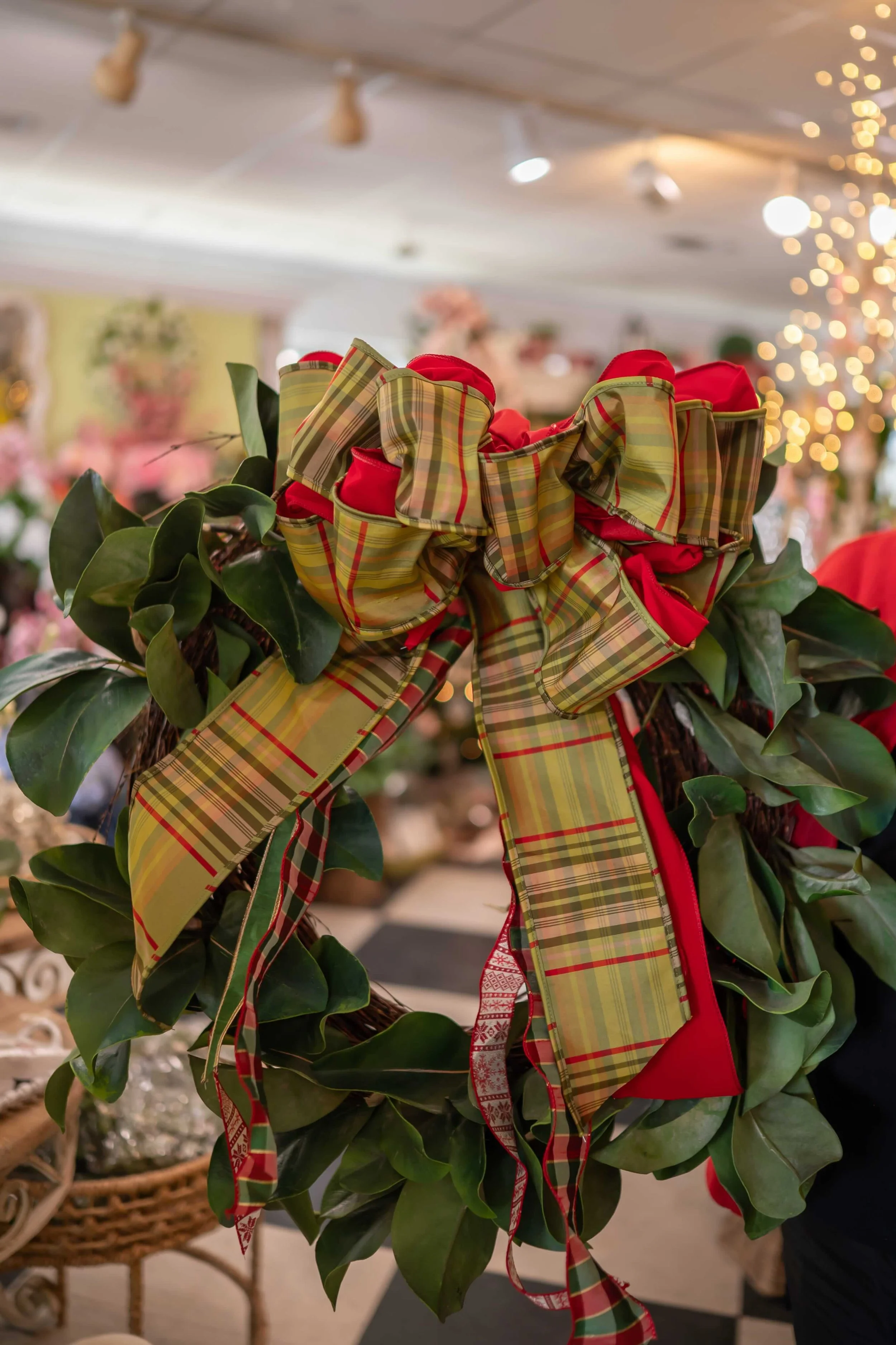 A Christmas wreath with red and green plaid ribbons and green leaves, decorated with bows, inside a store with blurry lights and holiday decorations in the background.
