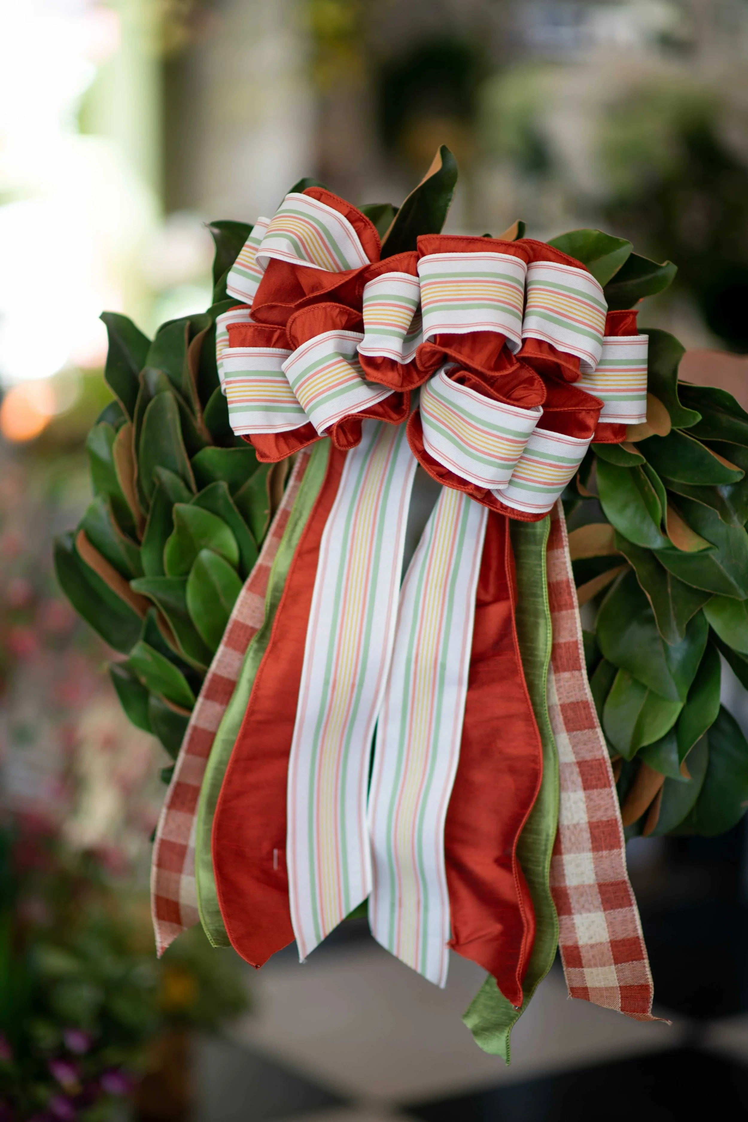 A decorative wreath with green leaves and red, white, and green striped ribbons, hanging outdoors.