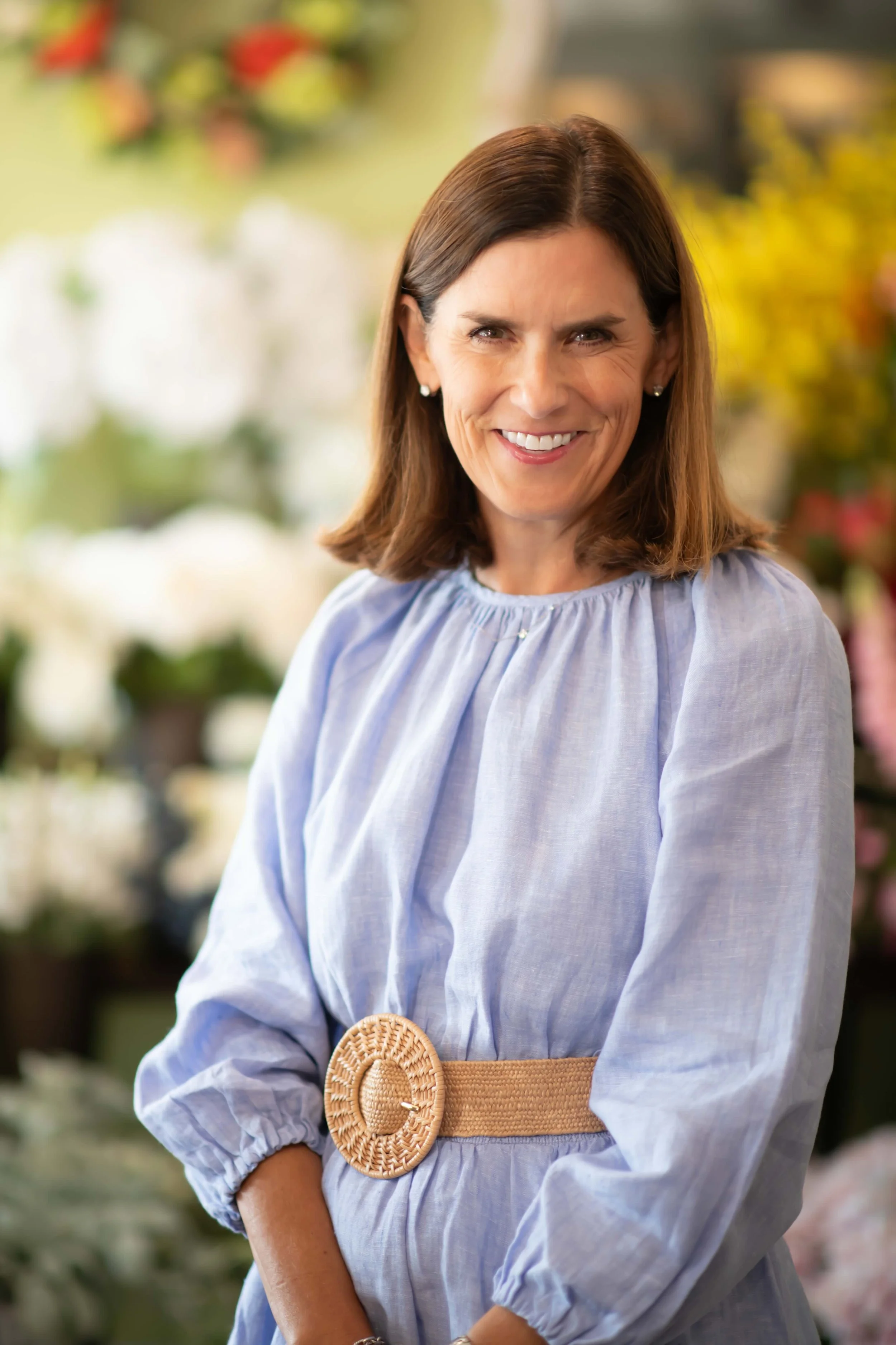 A woman with brown hair smiling, wearing a light blue dress with puffed sleeves and a tan woven belt with a round buckle, standing in a floral shop with blurred flowers in the background.