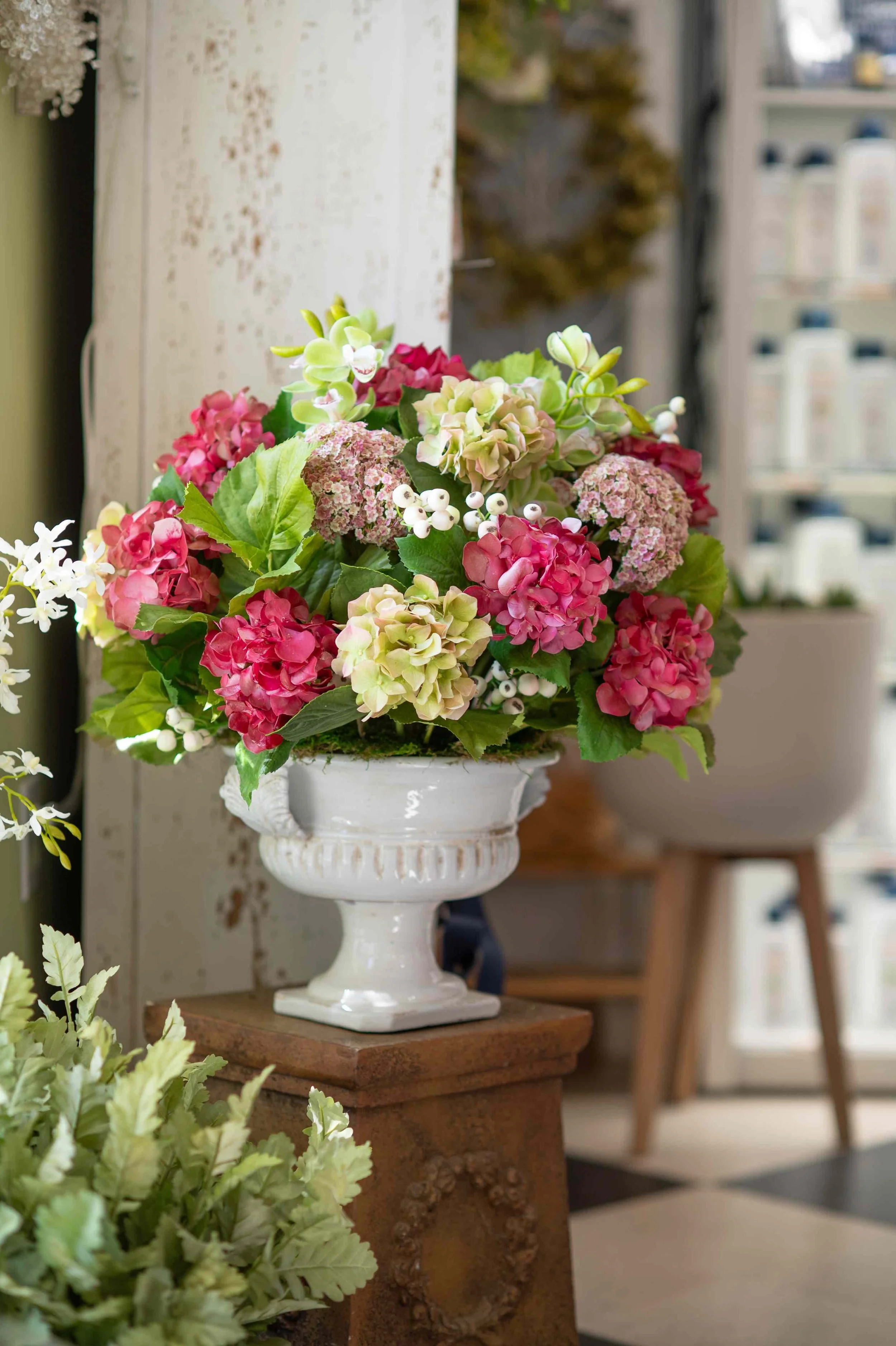 A bouquet of pink, white, and green flowers arranged in a white decorative vase, placed on a wooden pedestal in a store or shop setting.