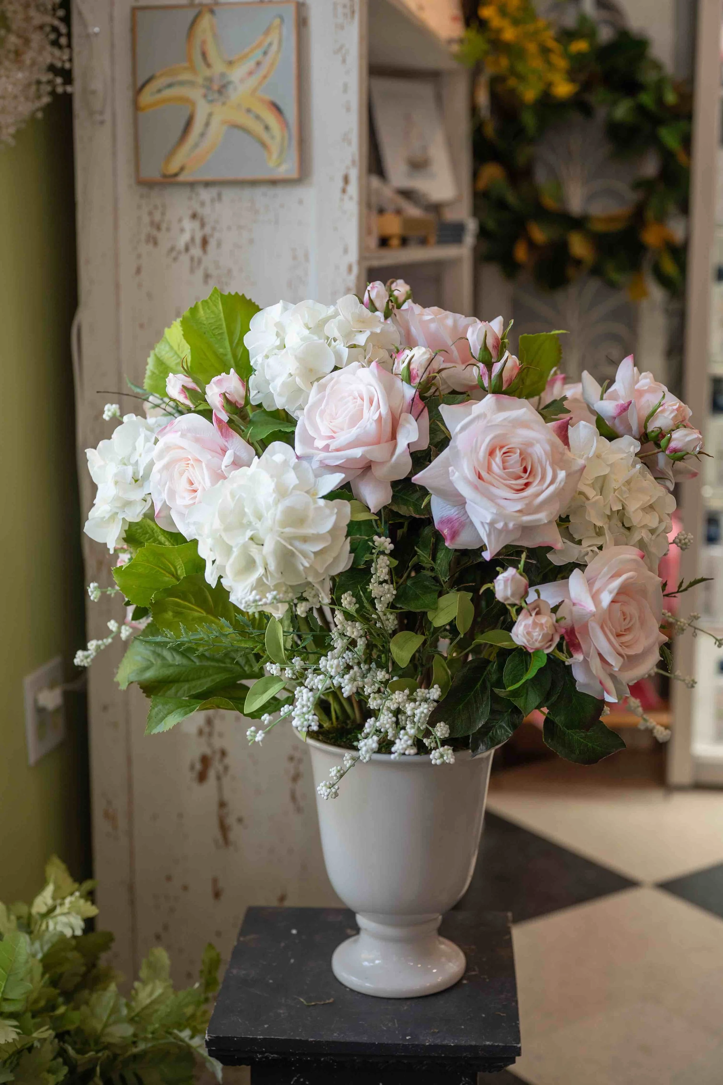 A bouquet of pink and white roses, white hydrangeas, and greenery arranged in a white vase, placed on a black stand, in a floral shop or display area.